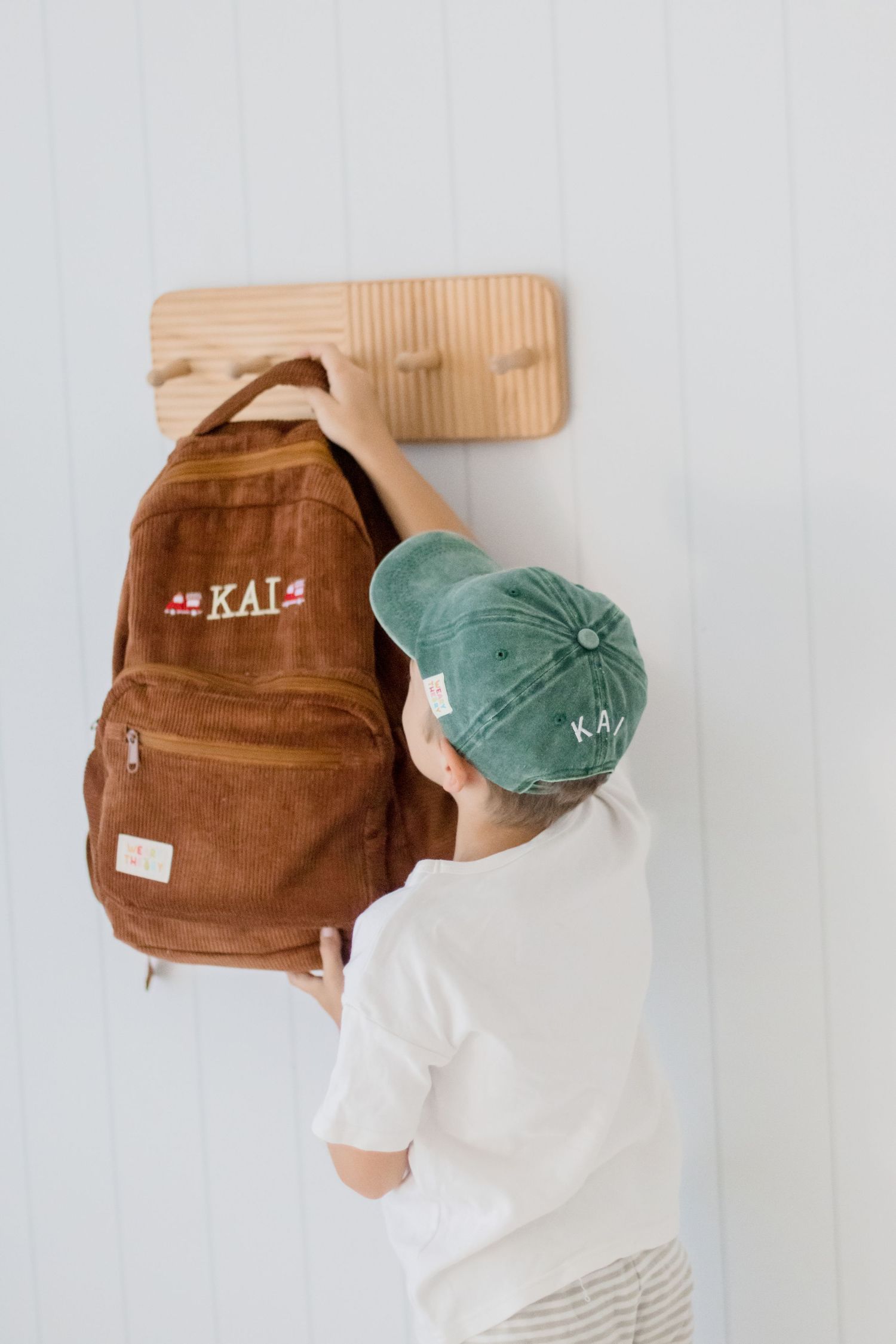 Brown backpack with text 'KAI' hanging on wooden wall hook with green cap.