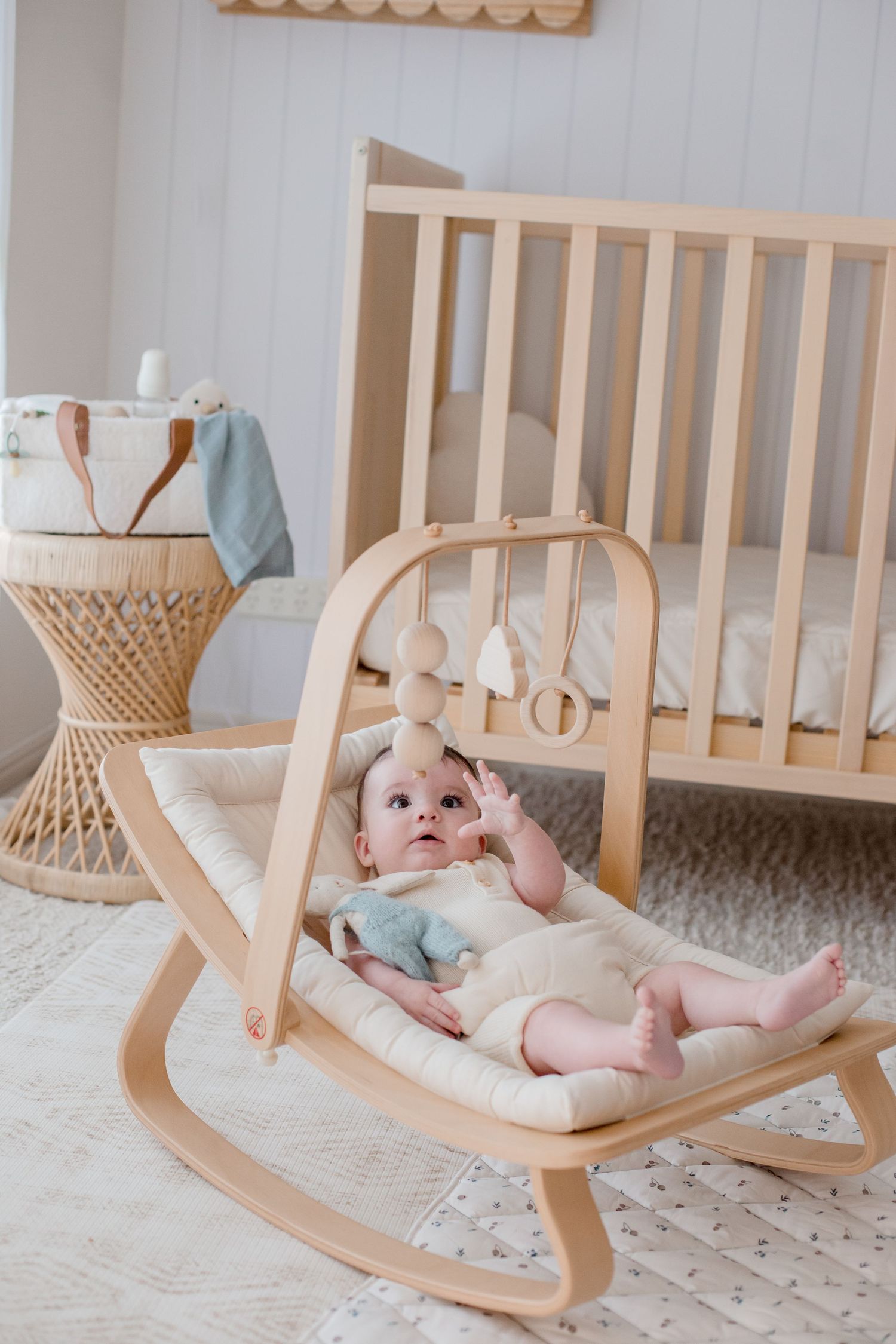 Baby relaxing on wooden rocker with hanging decorations in minimalist nursery.