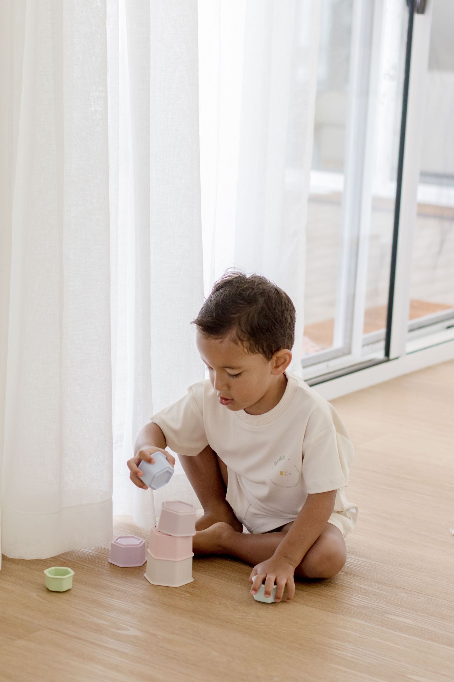 Child in white shirt playing with pastel stacking cups on wooden floor.