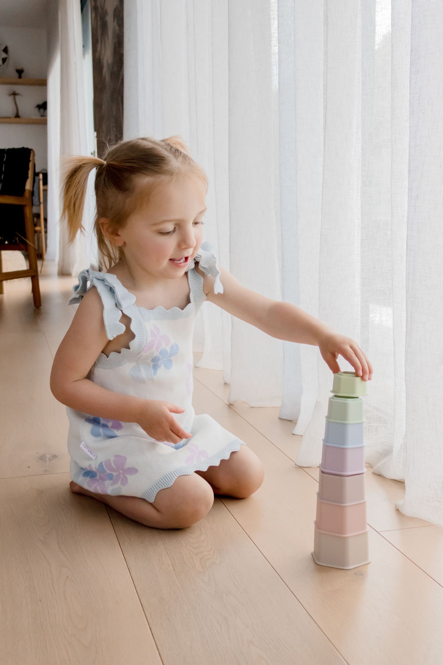 Child in floral dress stacking colorful pastel cups near window.