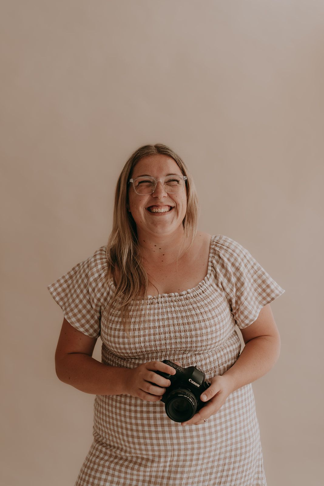 Photographer in gingham dress holding camera against neutral background.