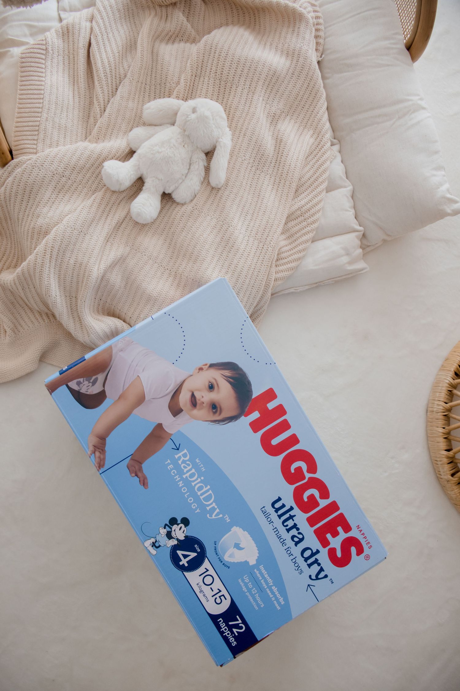 Box of Huggies diapers on white bedding with a plush bunny toy and ribbed beige blanket.