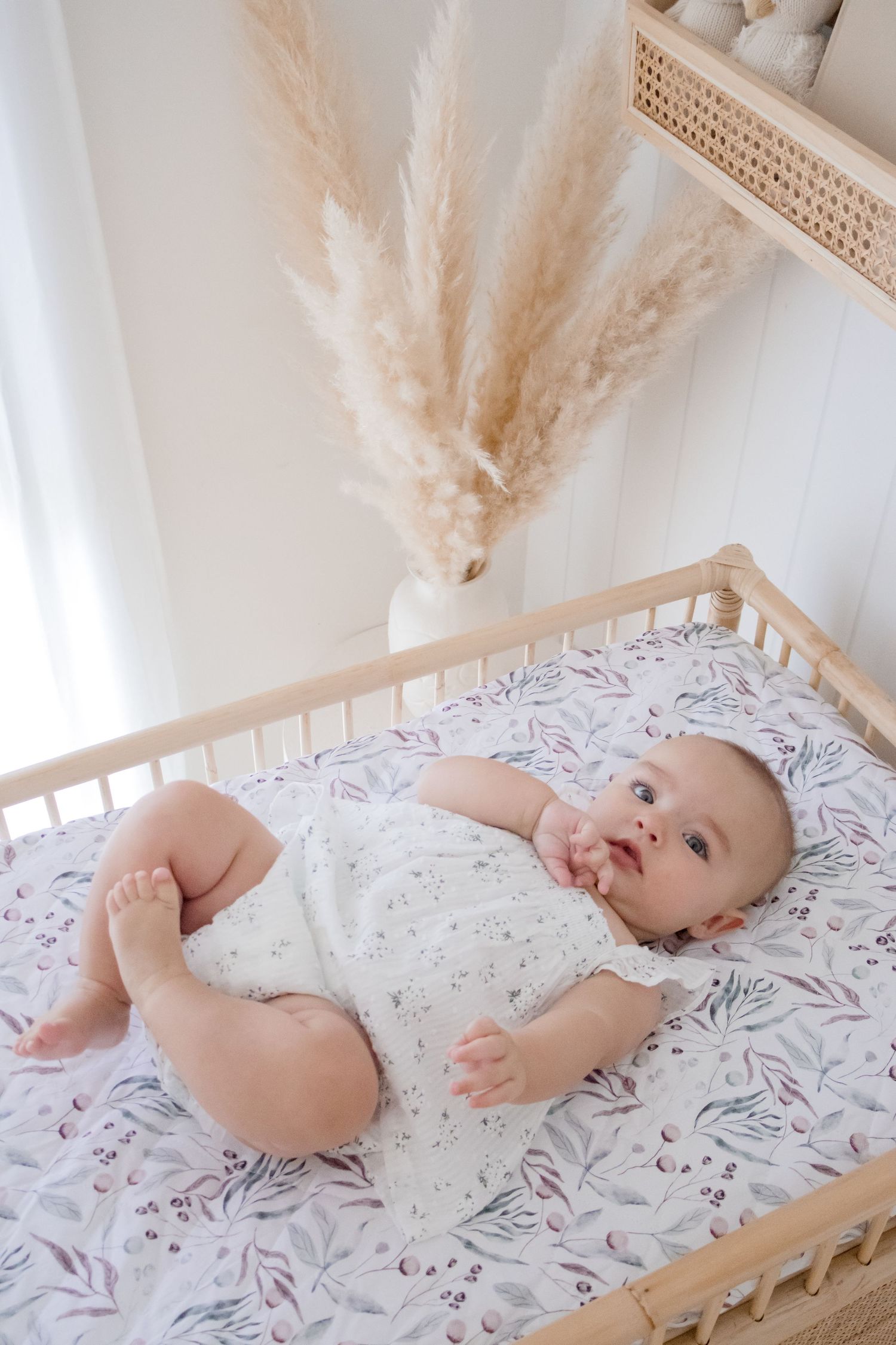 Baby lying on floral crib sheet with pampas grass decor in background.