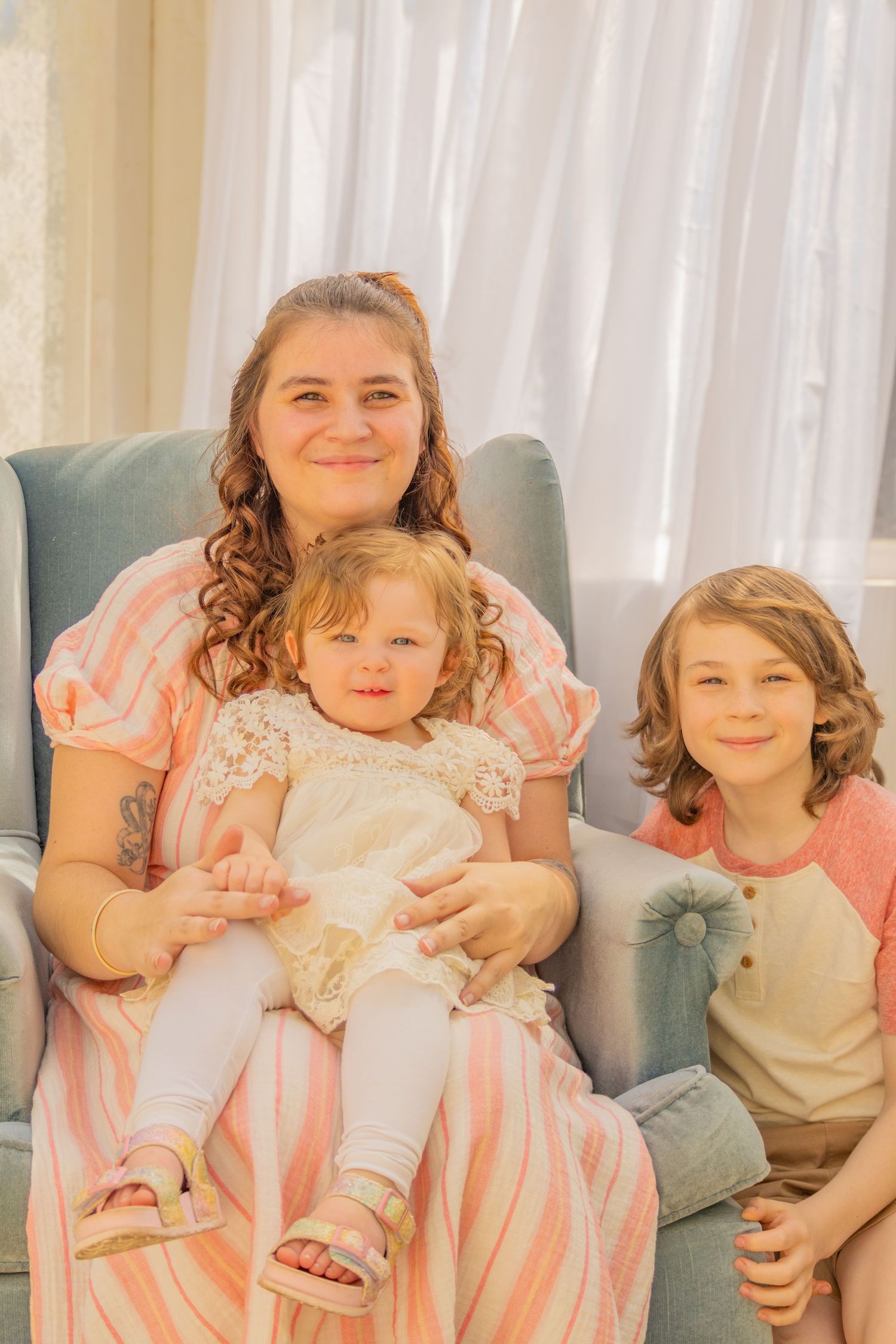 A family sits together on a blue armchair in soft lighting, wearing coordinating pink and coral outfits.