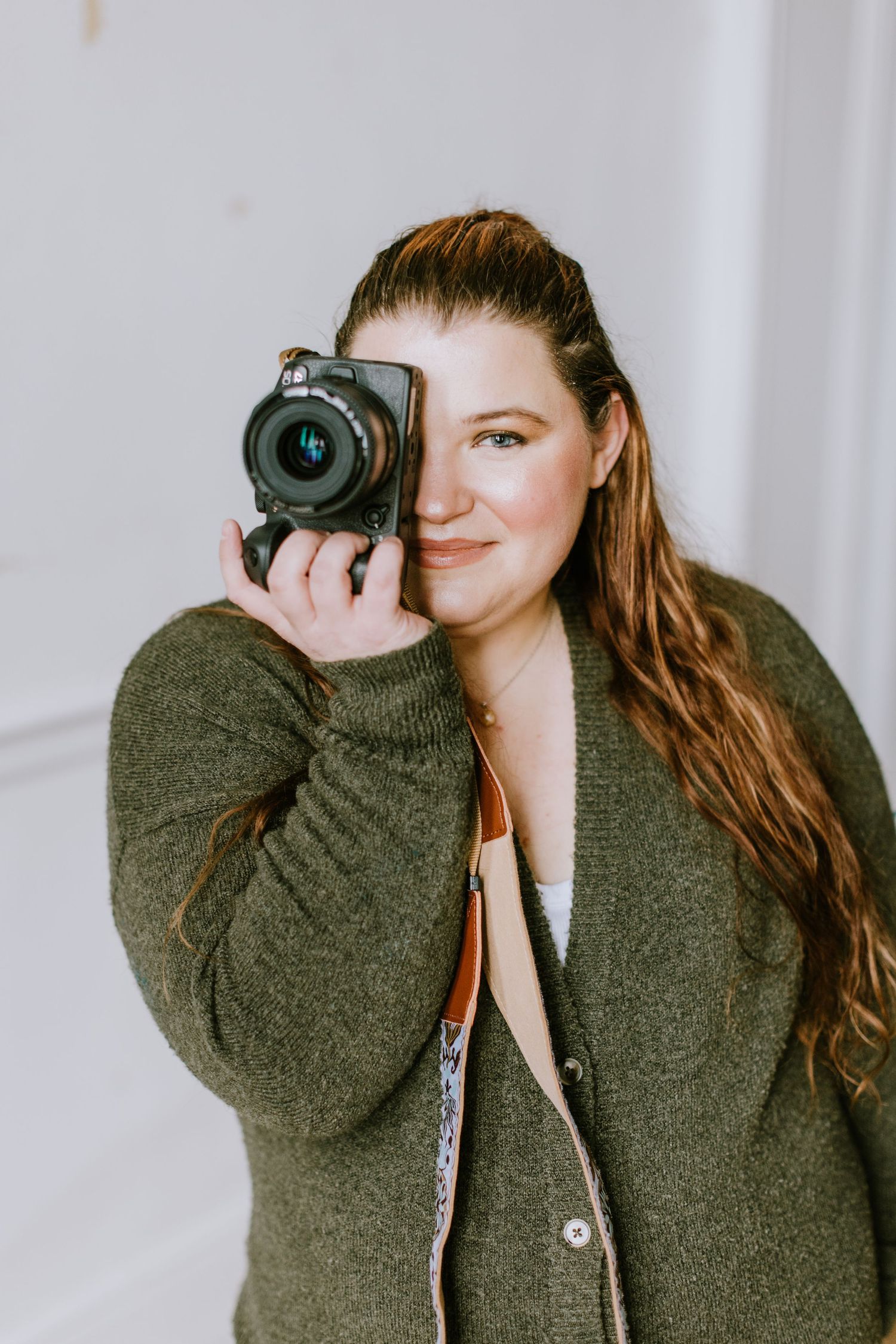 Person in green sweater holding camera in front of white background.