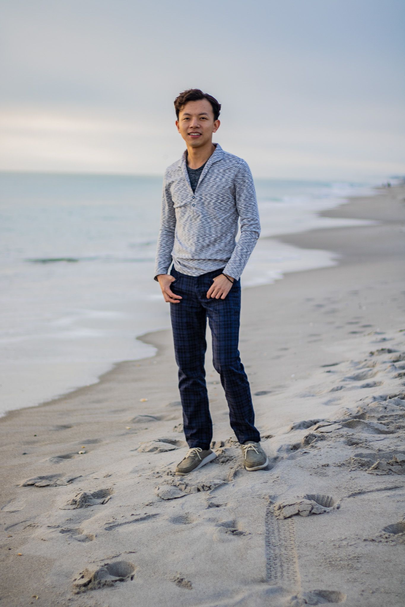 Person in casual sweater stands on beach at sunset with ocean waves in background.