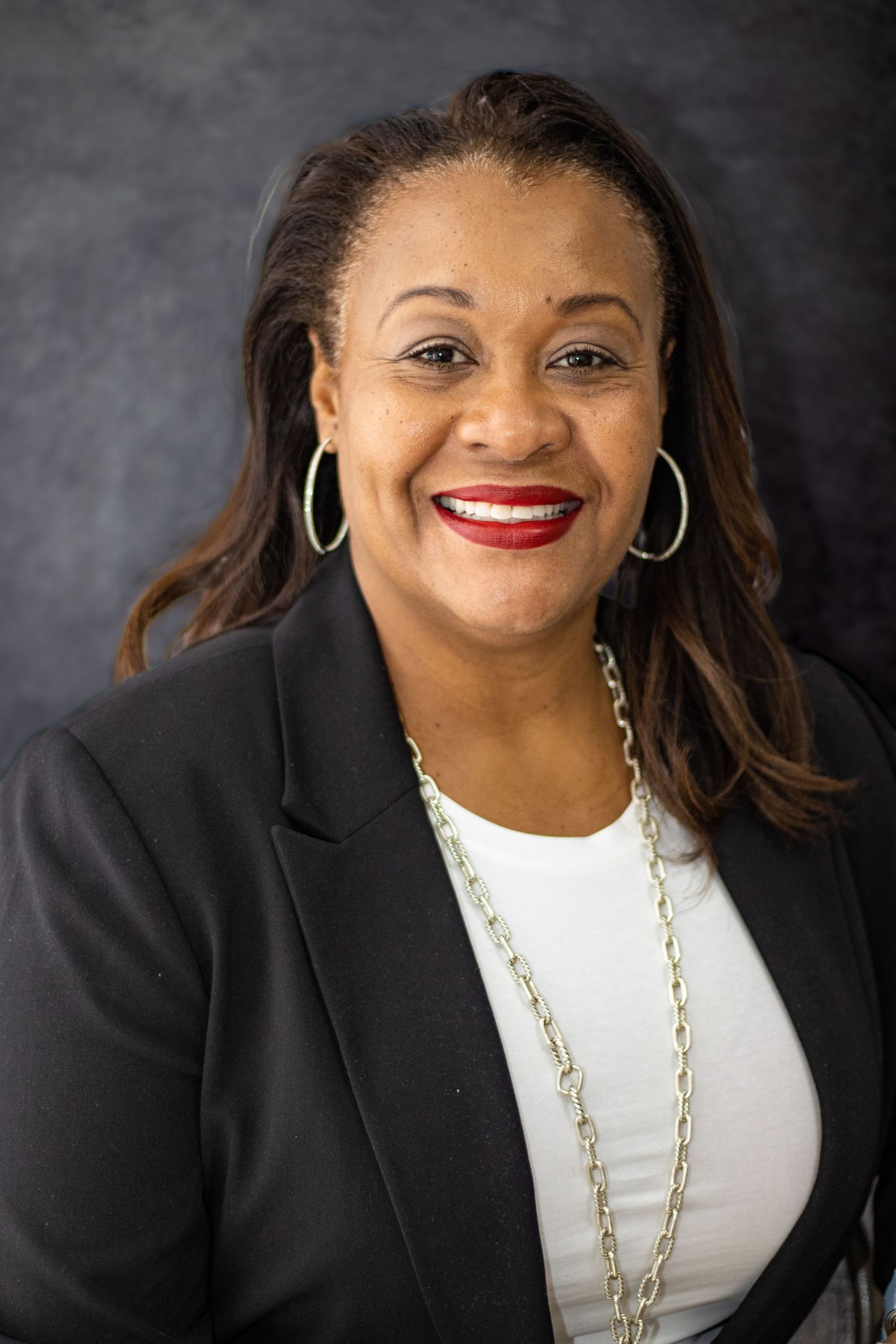 Professional portrait in black blazer and white top with silver necklace against gray backdrop.