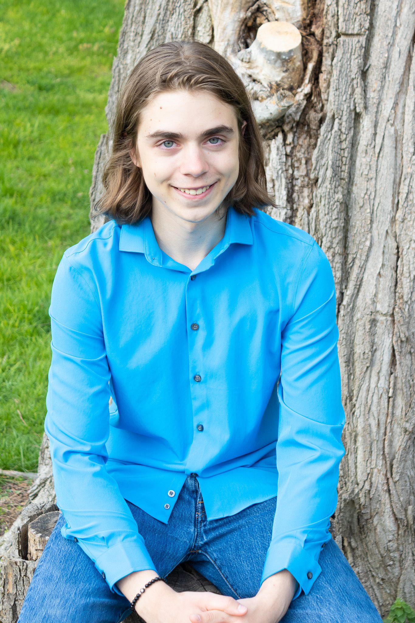 Student sits casually against a tree trunk wearing a bright blue button-down shirt and jeans on a sunny day.