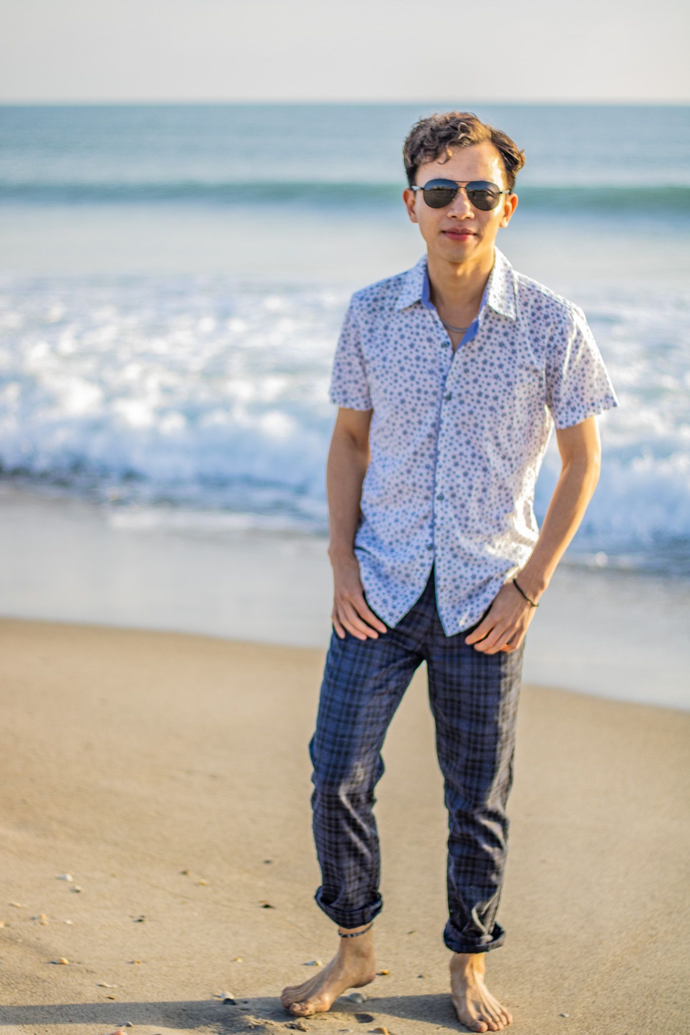 Man wearing sunglasses and floral shirt poses casually on sunny beach with ocean waves in background.