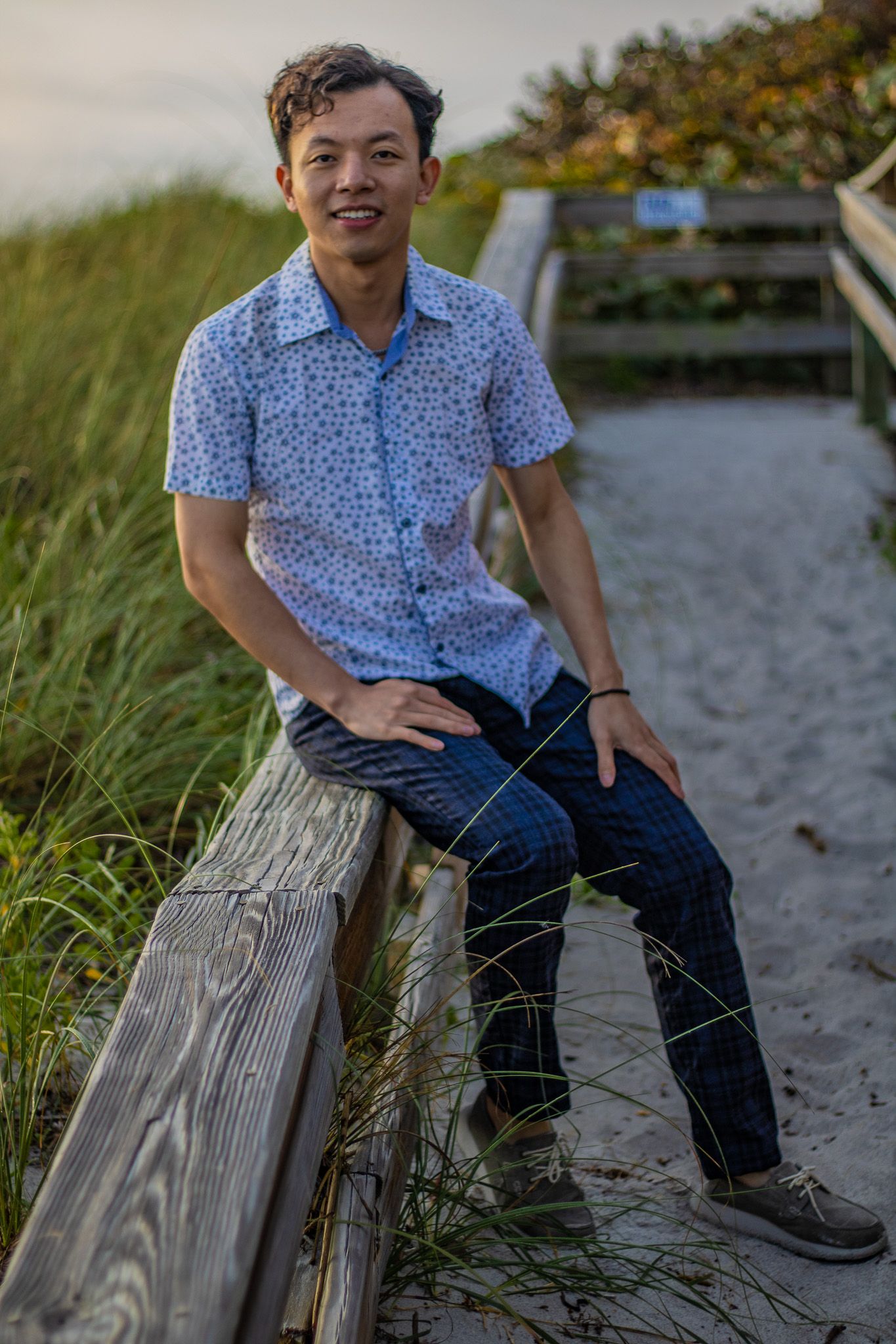 A casual portrait on a wooden beach walkway wearing a blue floral shirt and jeans at sunset.