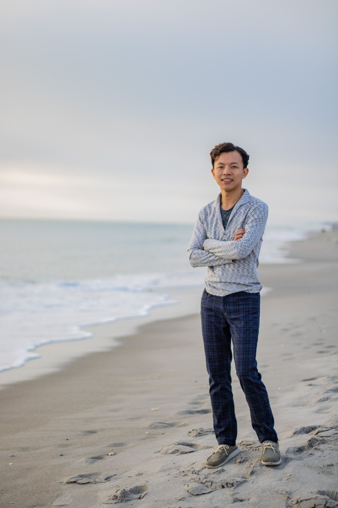 Figure stands with crossed arms on sandy beach during cloudy evening.