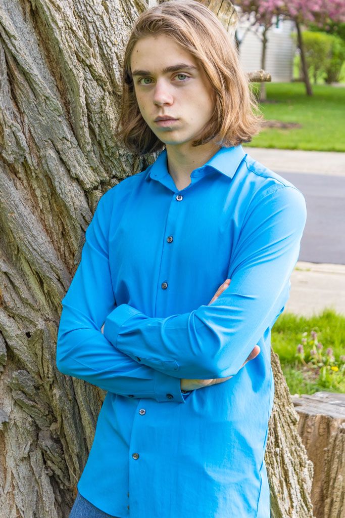 Person in bright blue shirt leans against large tree trunk with spring blossoms in background.