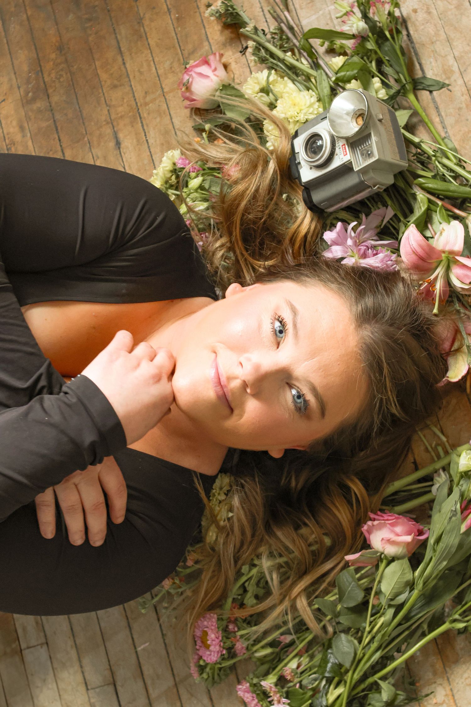Person in black dress lying on wooden floor surrounded by pink flowers and vintage camera.