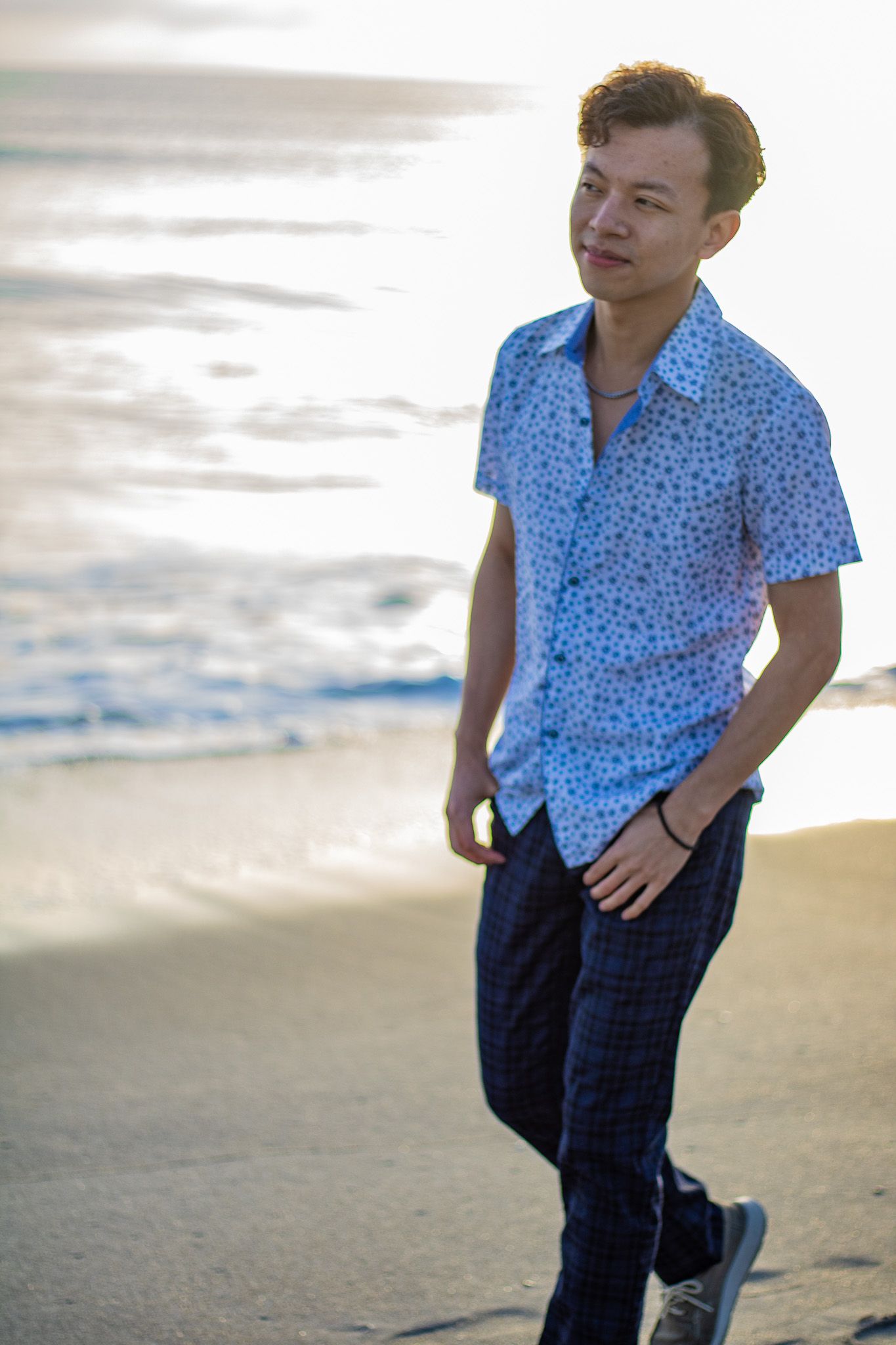 Person in blue patterned shirt poses casually on beach at sunset.
