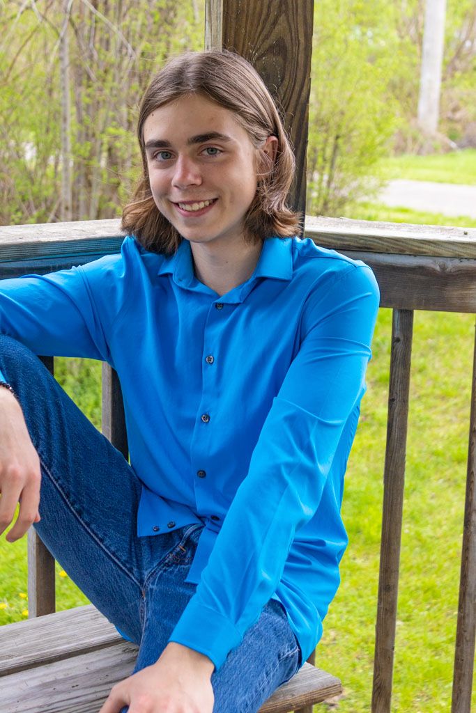 Person in vibrant blue shirt sits relaxed on wooden deck railing with green foliage in background.