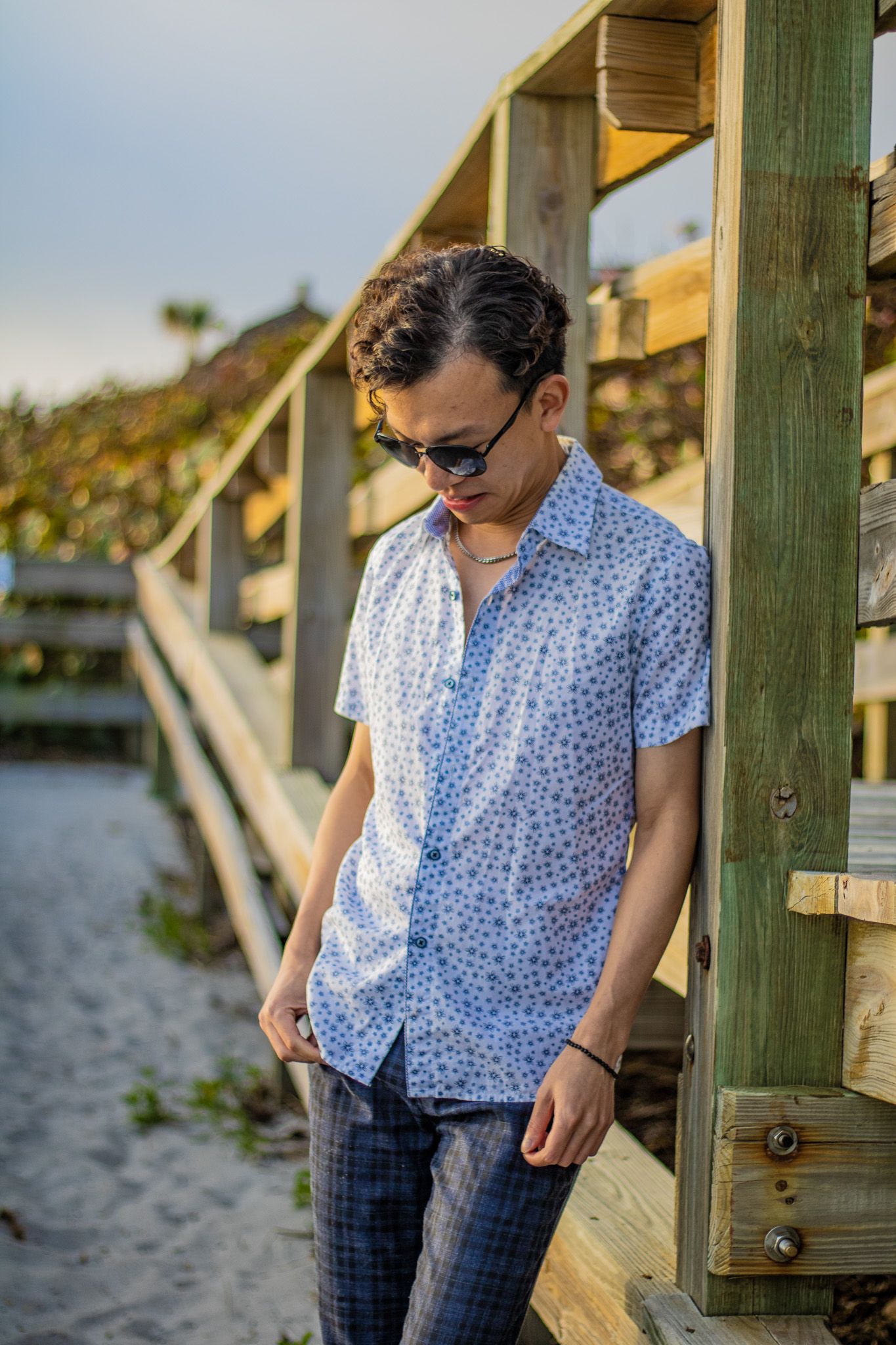 Fashion portrait with sunglasses and blue patterned shirt on sandy coastal boardwalk.