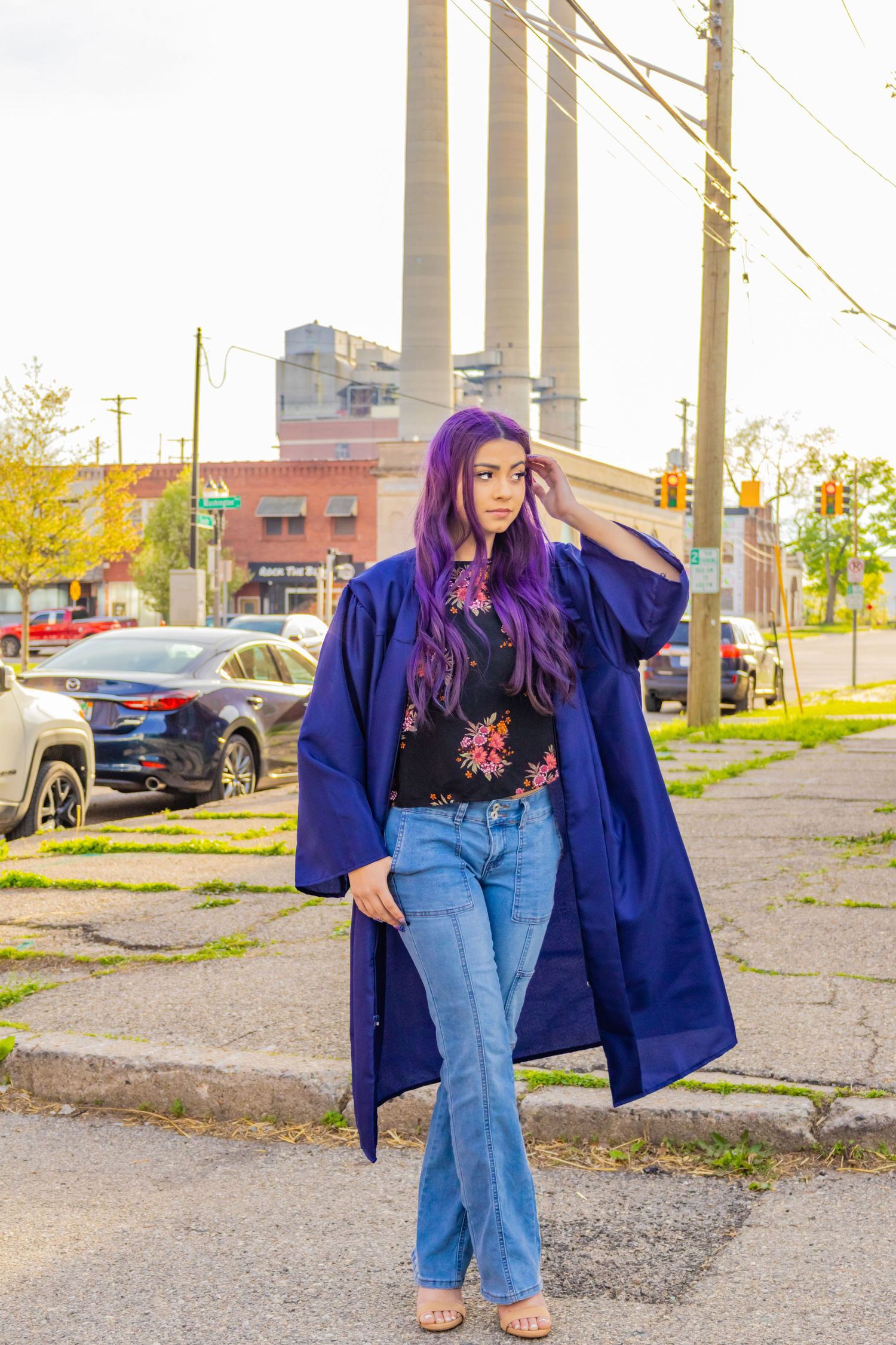A figure with purple hair wearing a navy graduation gown, floral top and jeans poses in an industrial urban setting.