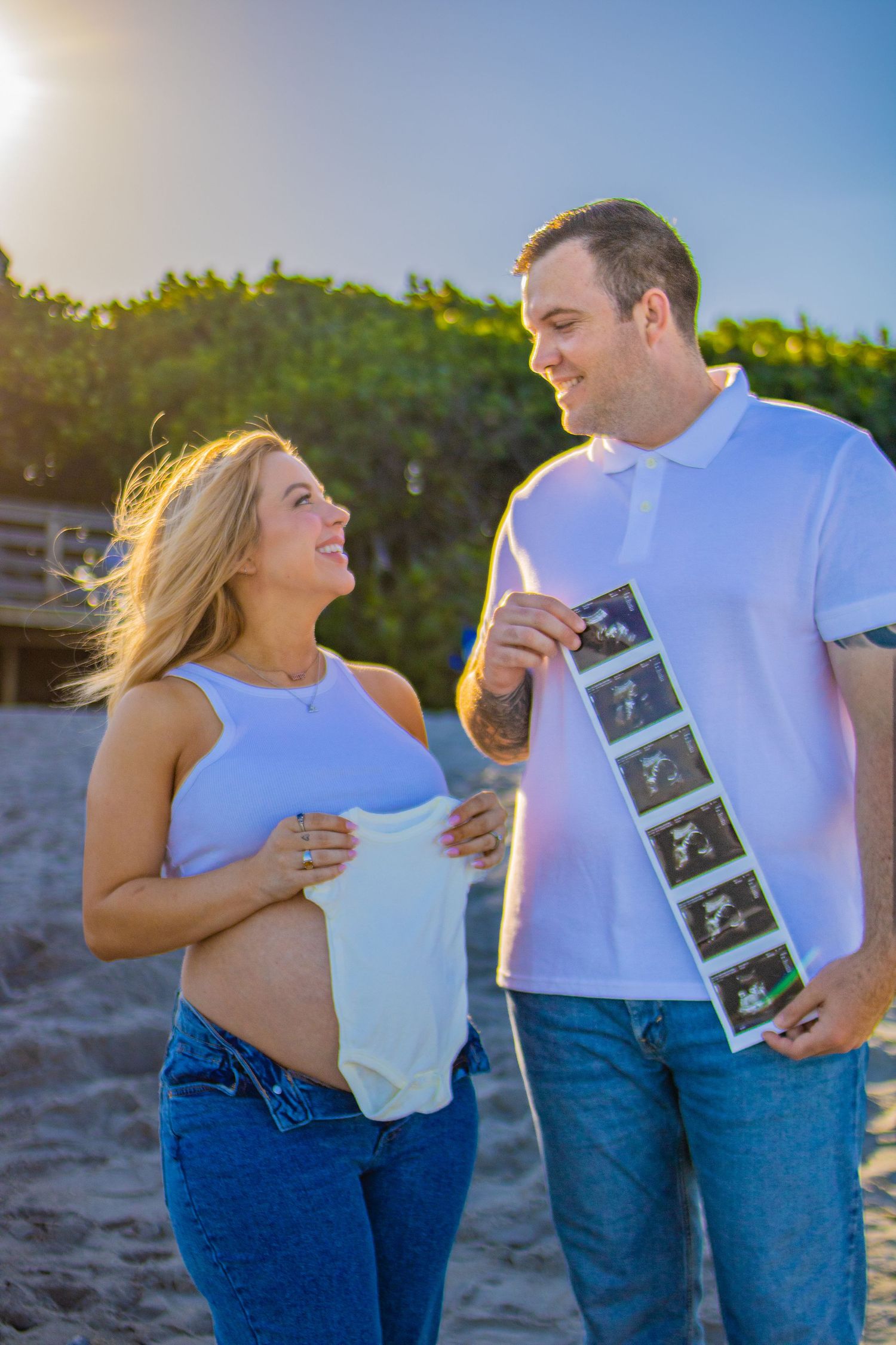 Expectant couple on beach share ultrasound photos during sunset photoshoot.