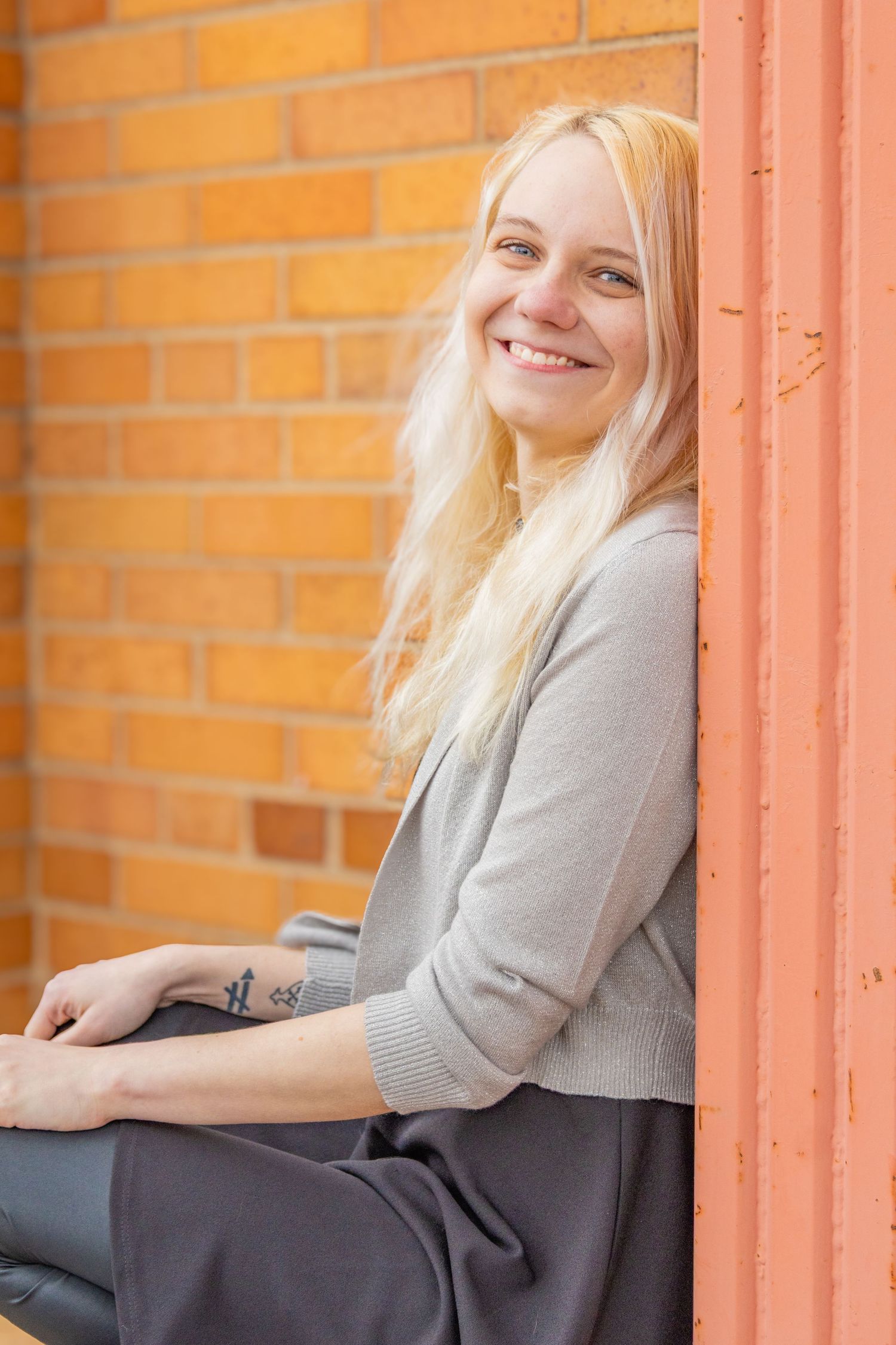A person with long blonde hair wearing a gray sweater sits casually against an orange brick wall.