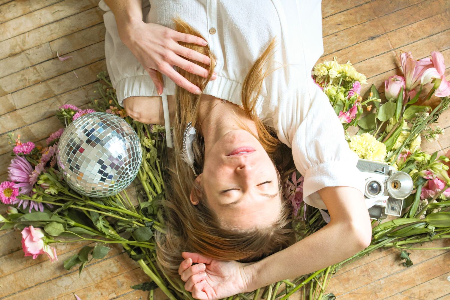 Person lying on wooden floor with disco ball and scattered pink flowers in dreamy portrait.