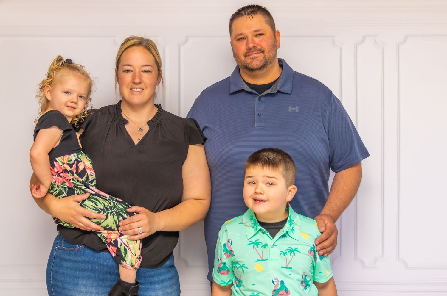 Family poses for a casual portrait against a white wall in casual clothing.