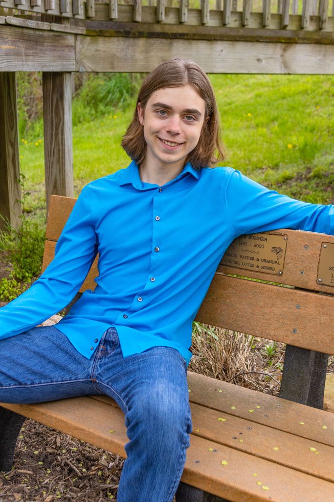 Person in bright blue shirt sits casually on wooden park bench with green grass background.