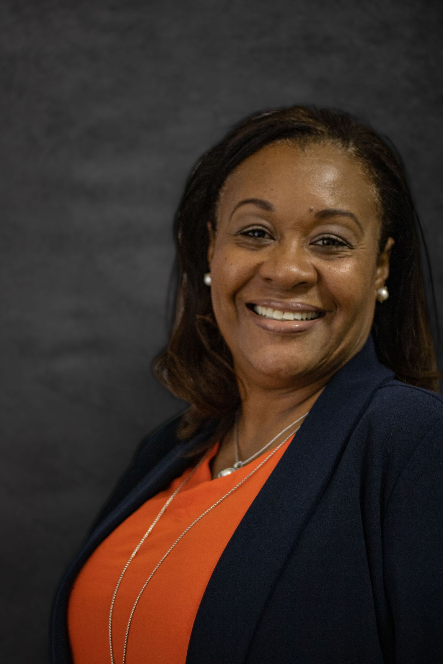 Professional headshot of person smiling warmly in orange top and navy blazer against dark background.