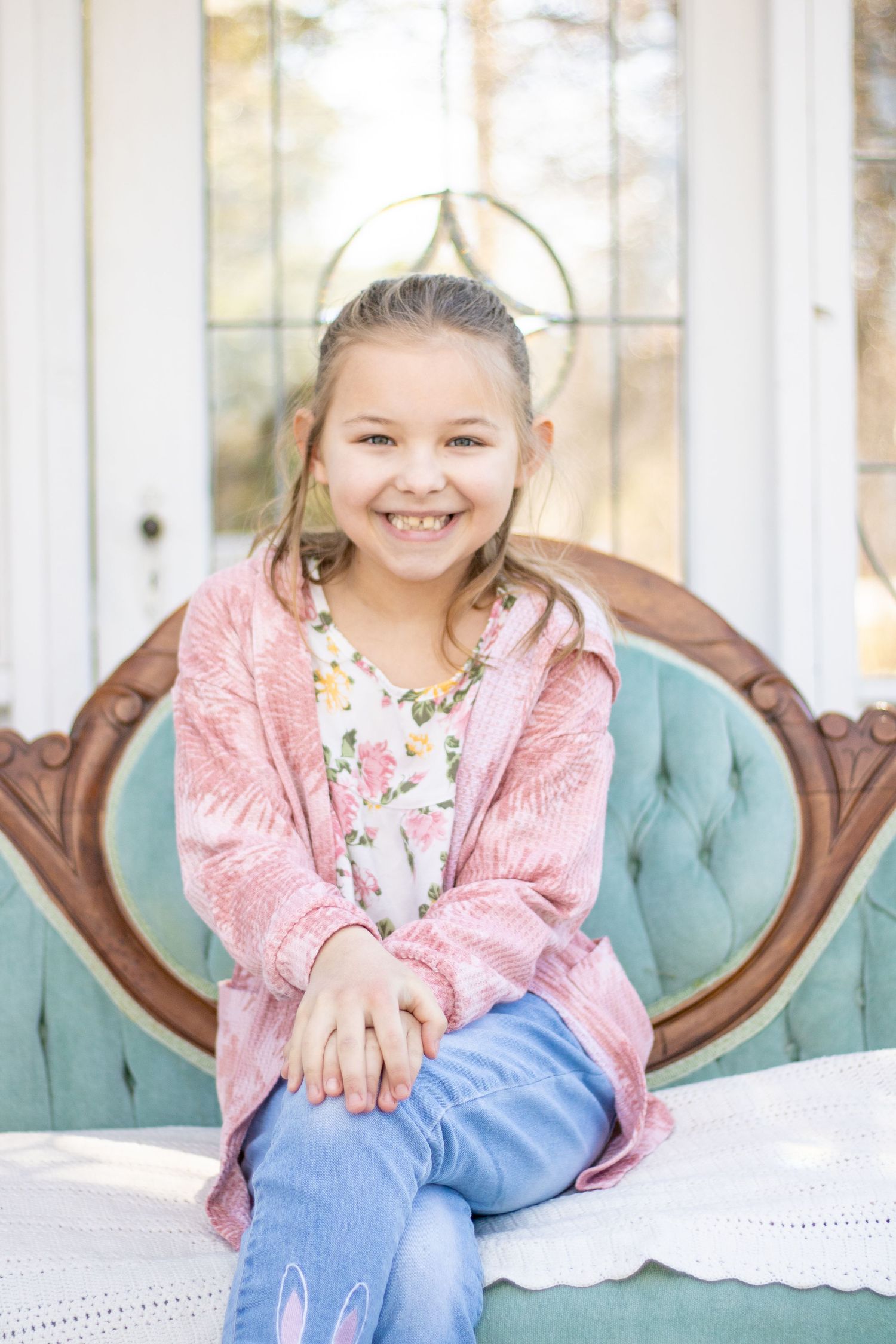 Child wearing pink cardigan and floral top sits on teal tufted couch.