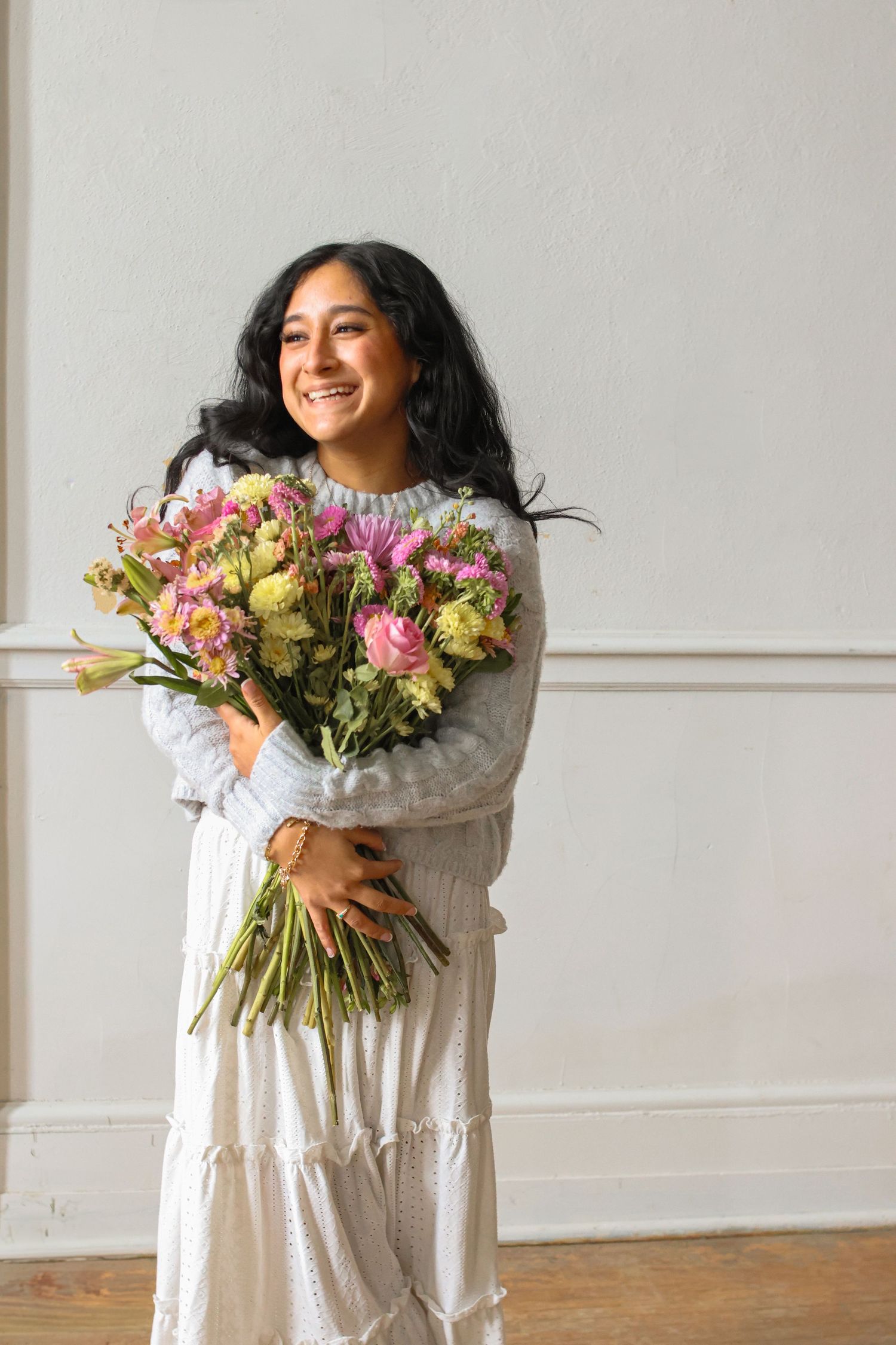 Person in white dress holding colorful spring bouquet with pink and yellow flowers against white wall.