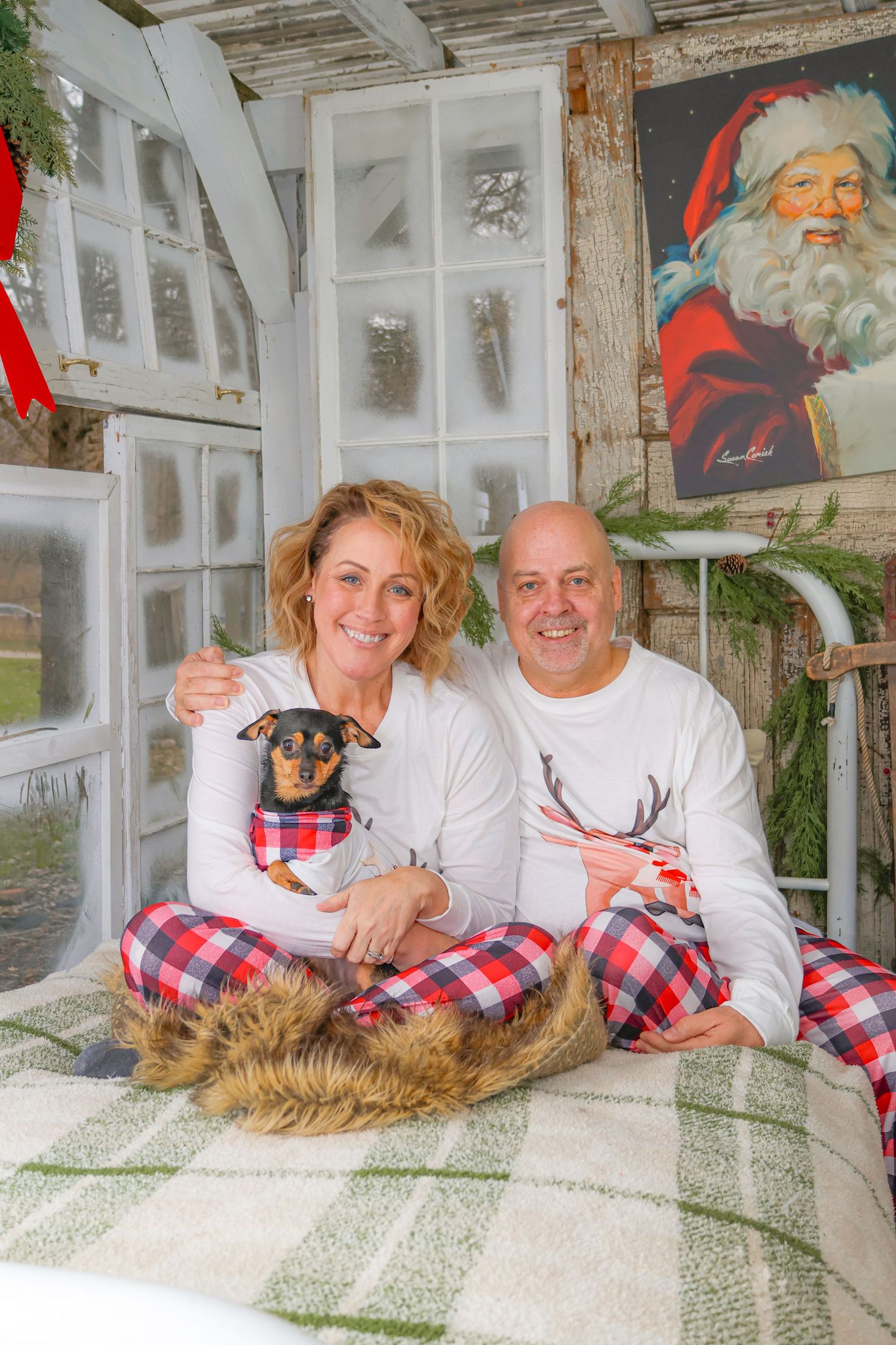 Couple in matching plaid pajamas poses with their small dog in front of a Santa Claus decoration.