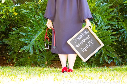 Person in graduation gown holding chalkboard sign and red shoes against green foliage.