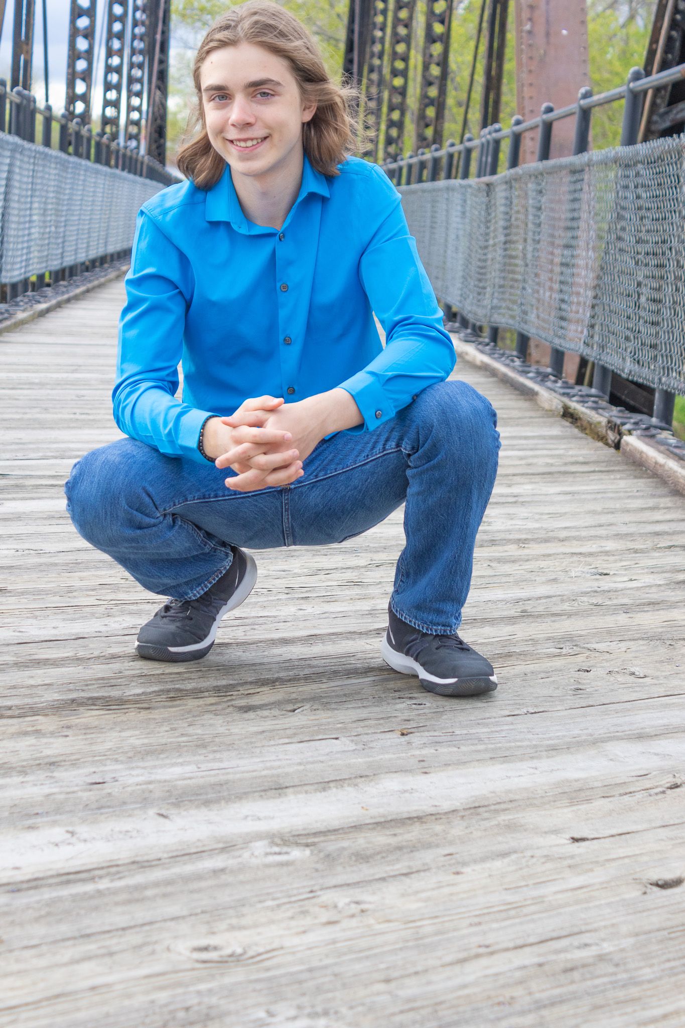 Person in bright blue shirt and jeans crouches on wooden bridge.