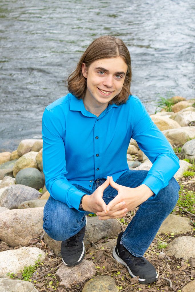 Person in blue button-down shirt crouches on rocky riverbank with flowing water behind.
