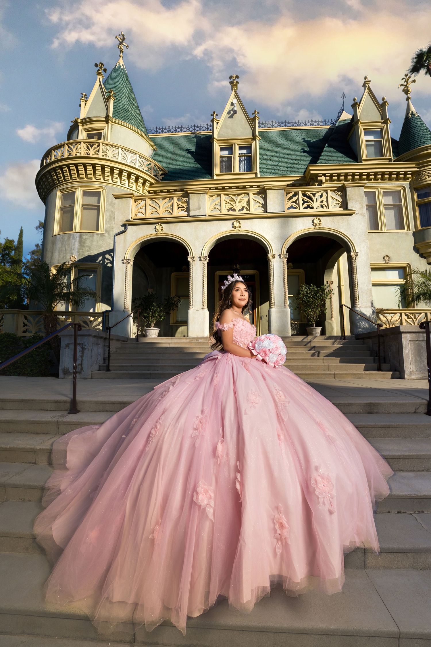 A person in a voluminous pink princess ball gown poses on the steps of an ornate Victorian mansion with turrets.