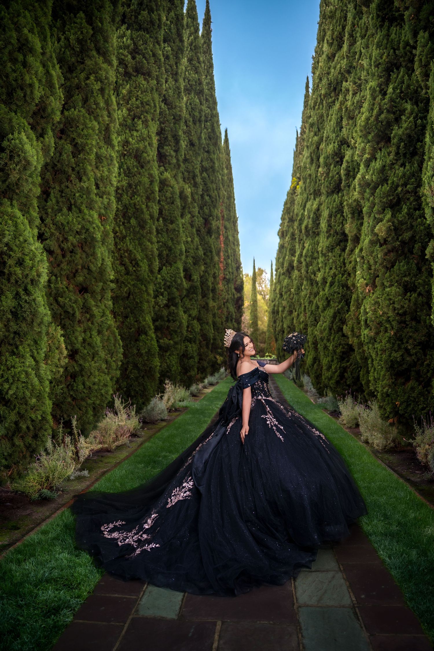 Ornate black gown with flowing train photographed along cypress tree-lined path under blue sky.