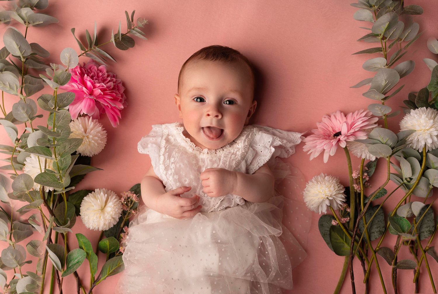 A baby in a white lace dress lies on a pink background surrounded by pink and white flowers.