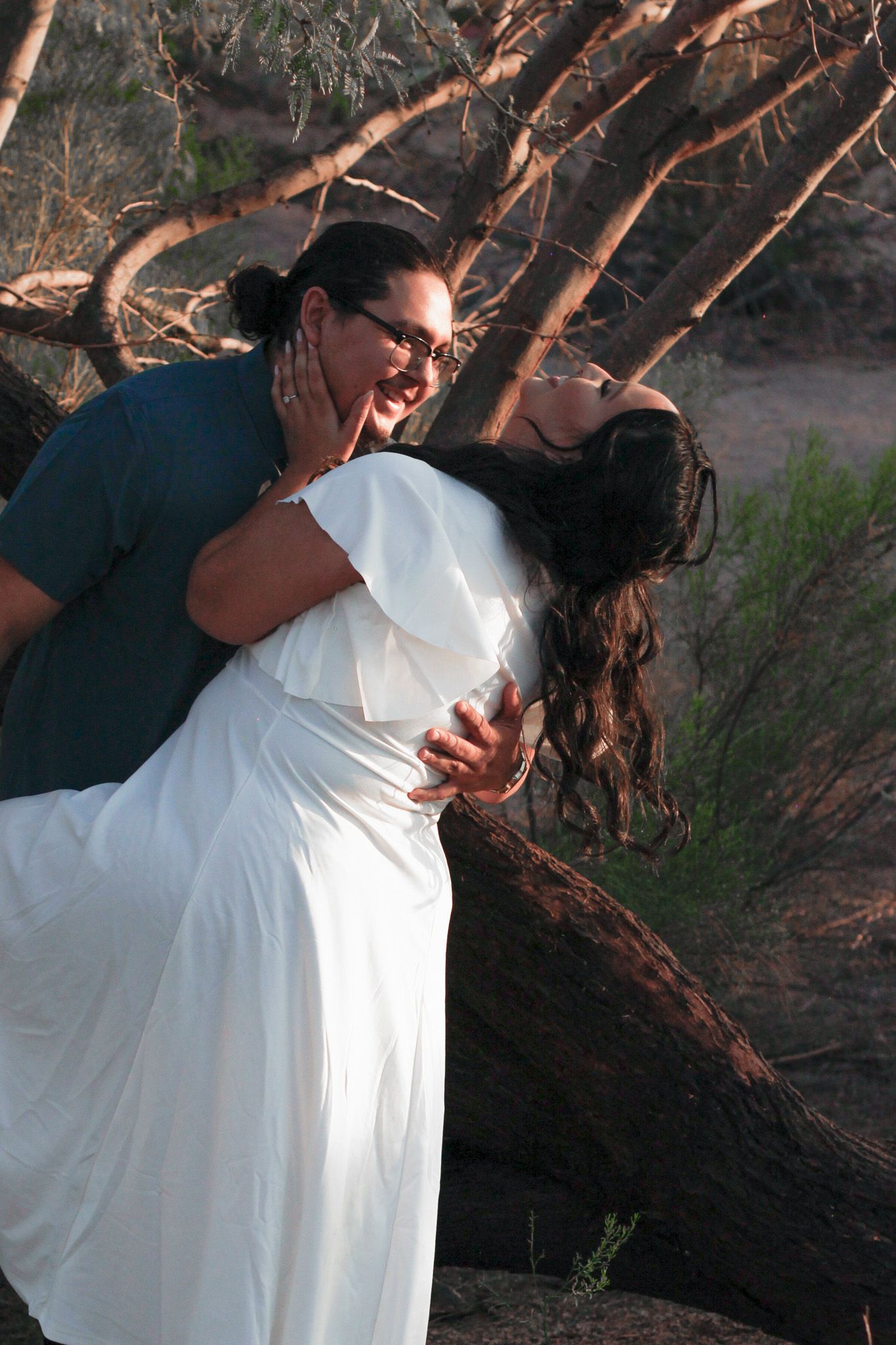 A person in glasses wearing dark clothing dips someone in a white dress against a backdrop of tree branches at sunset.