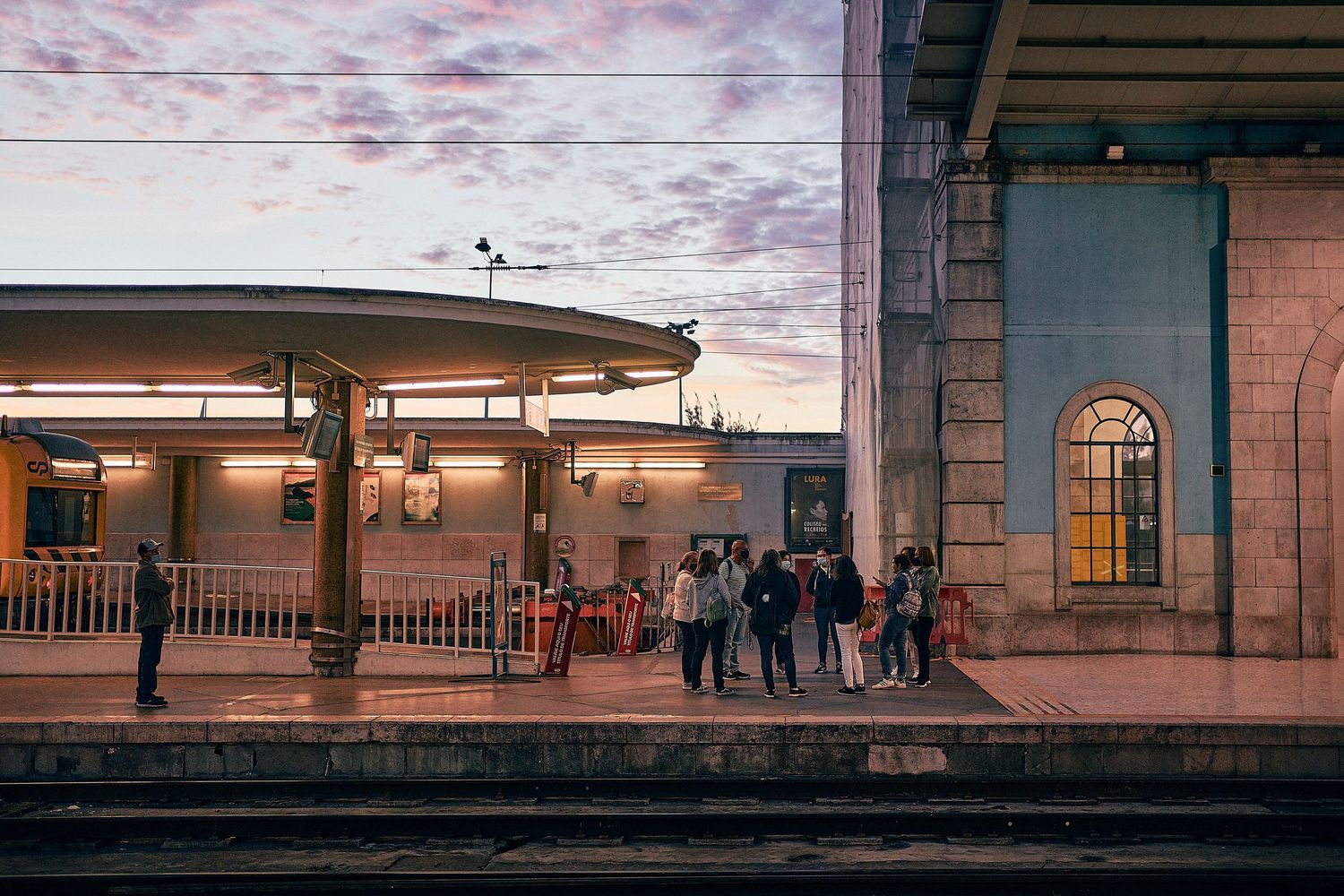 Passengers wait at a train station platform during sunset with pink clouds and vintage architecture.