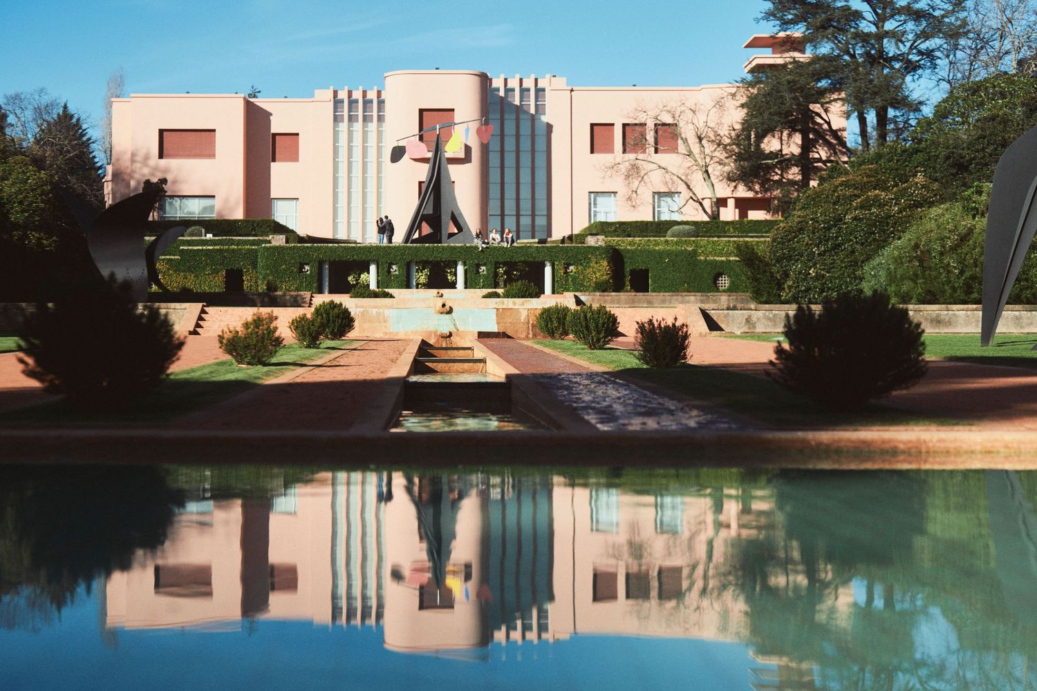 Art deco style pink mansion with reflecting pool and landscaped gardens shows symmetrical architectural design.