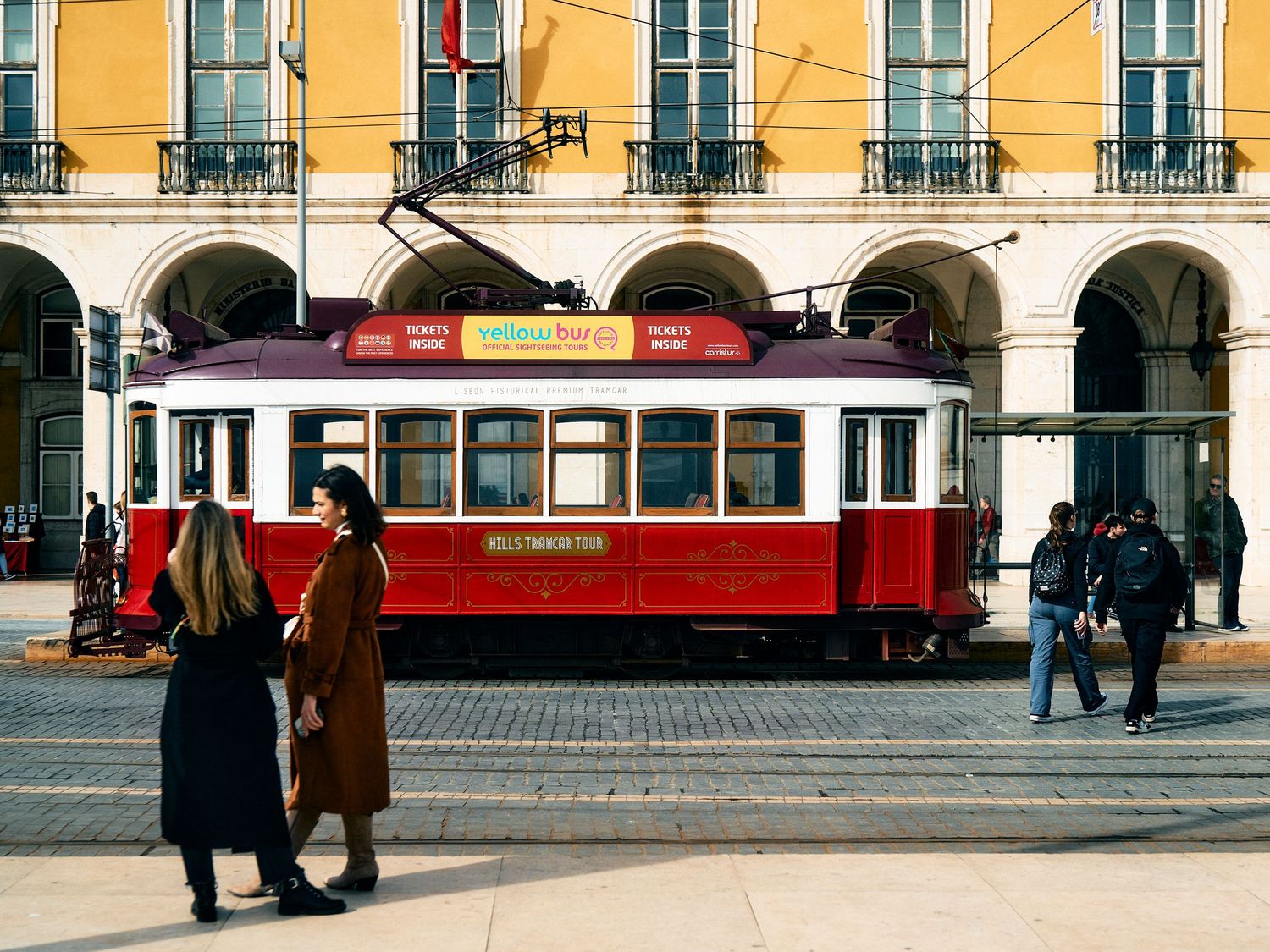 Red and white vintage tram on a street with arched columns and pedestrians in front of a yellow building in Lisbon.