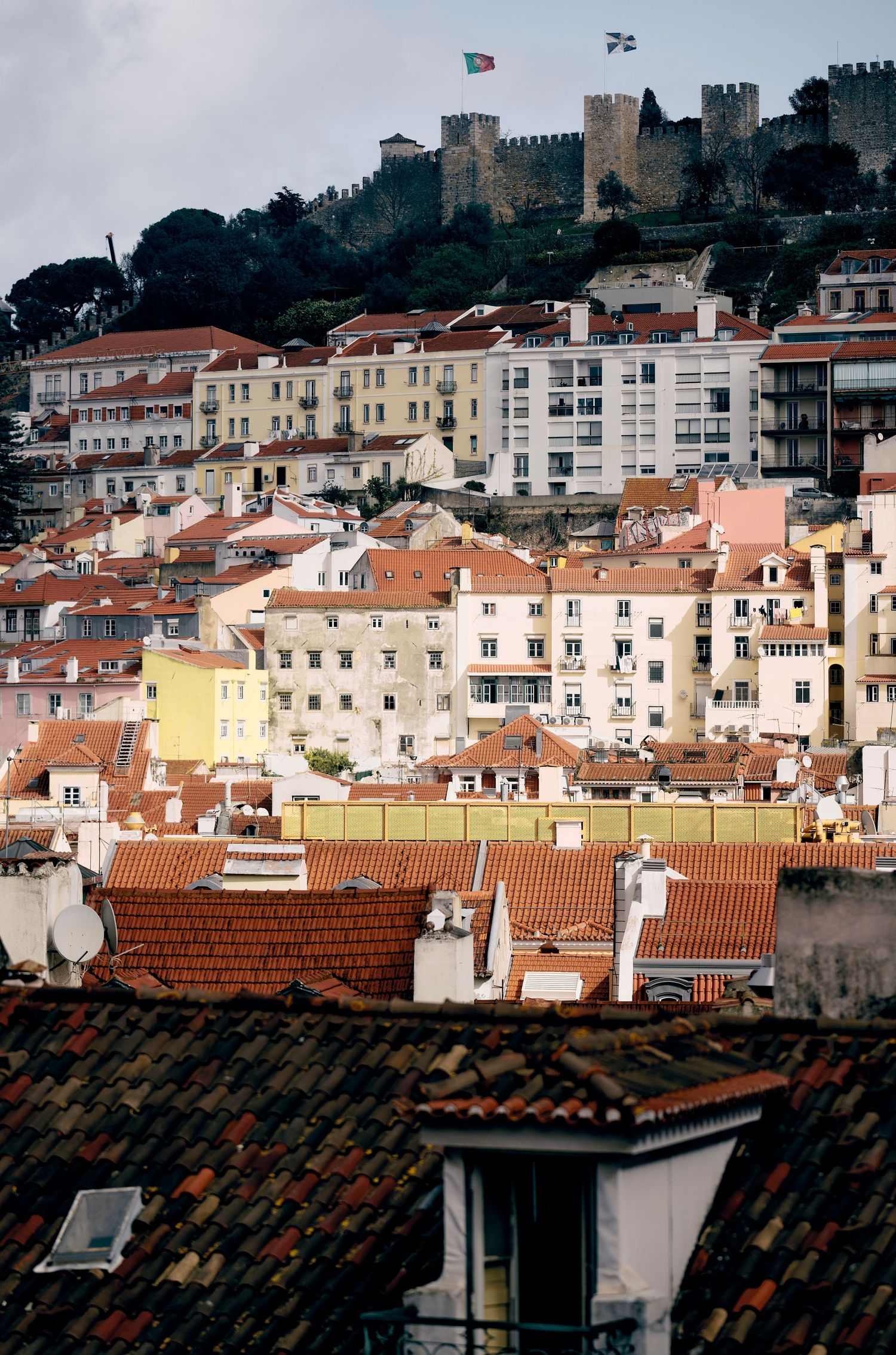 View of historic Lisbon cityscape with terracotta rooftops and hilltop Sao Jorge Castle overlooking dense urban buildings.