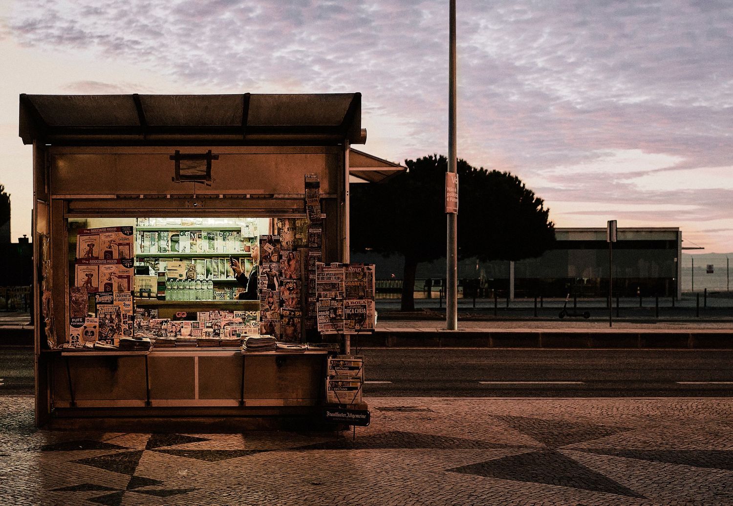 A small newspaper kiosk illuminated at dusk with a pink sky behind it.