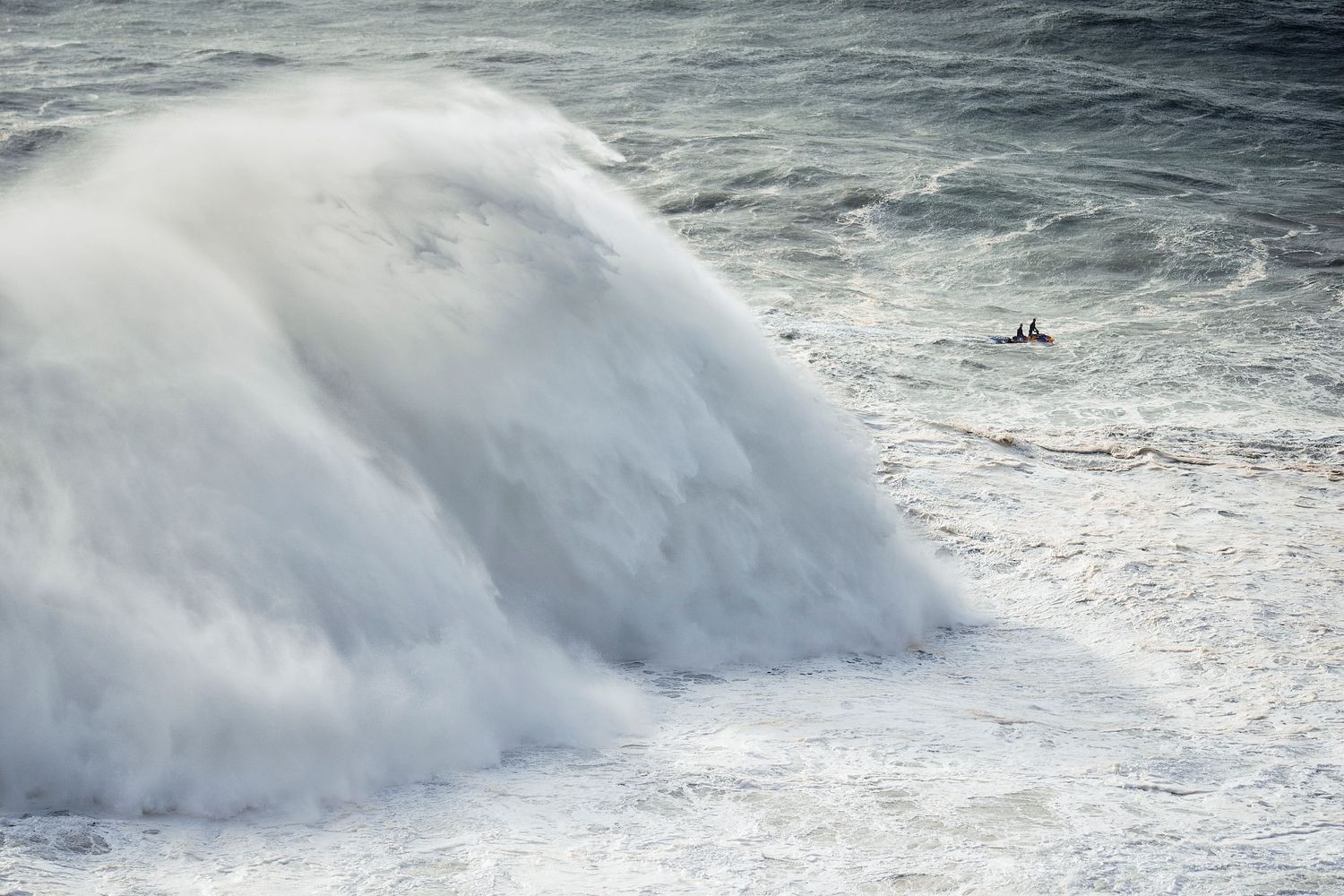 A massive ocean wave crashes with explosive force, creating a powerful white spray over dark churning waters.