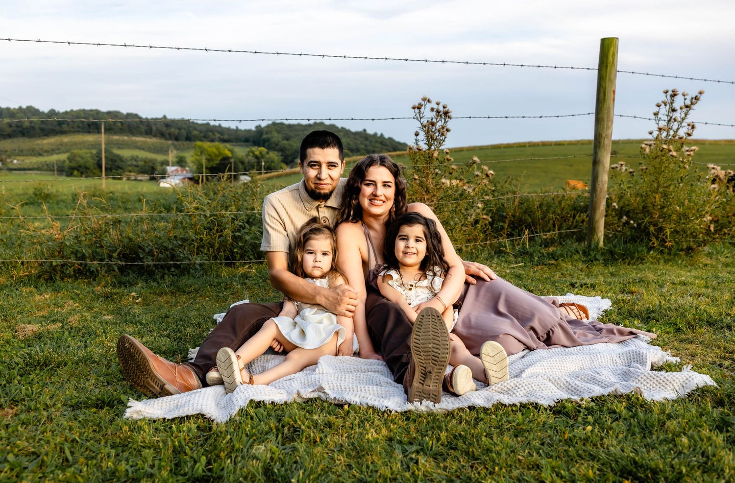 Family of four relaxing together on blanket in picturesque countryside setting during golden hour.