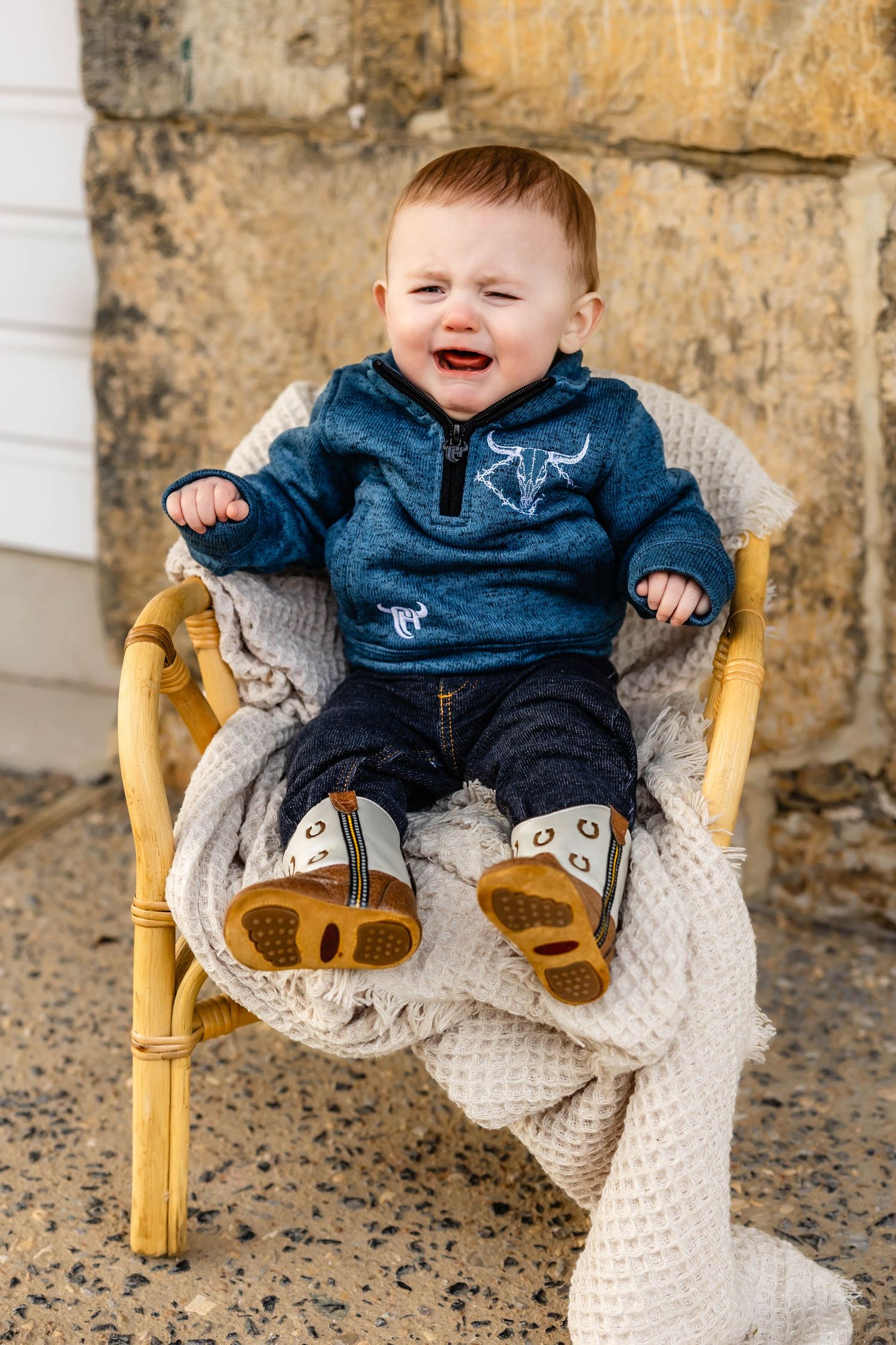 Happy baby sitting in yellow chair with knit blanket wearing blue hoodie.