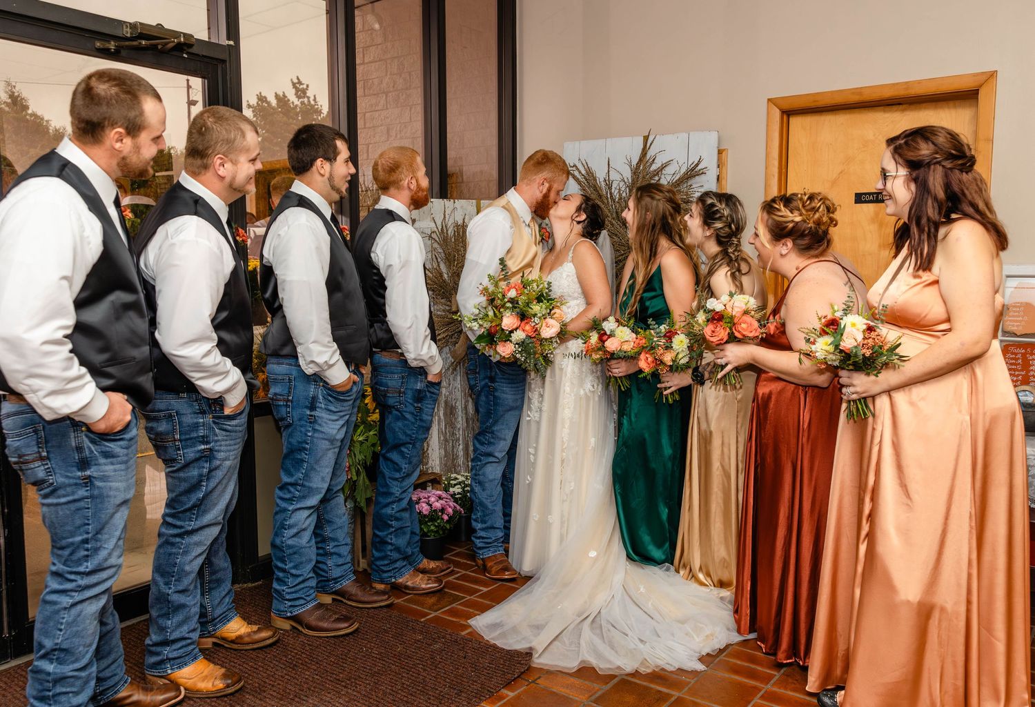 Wedding party lined up with groomsmen in jeans and vests facing bridesmaids in jewel-toned dresses holding bouquets.