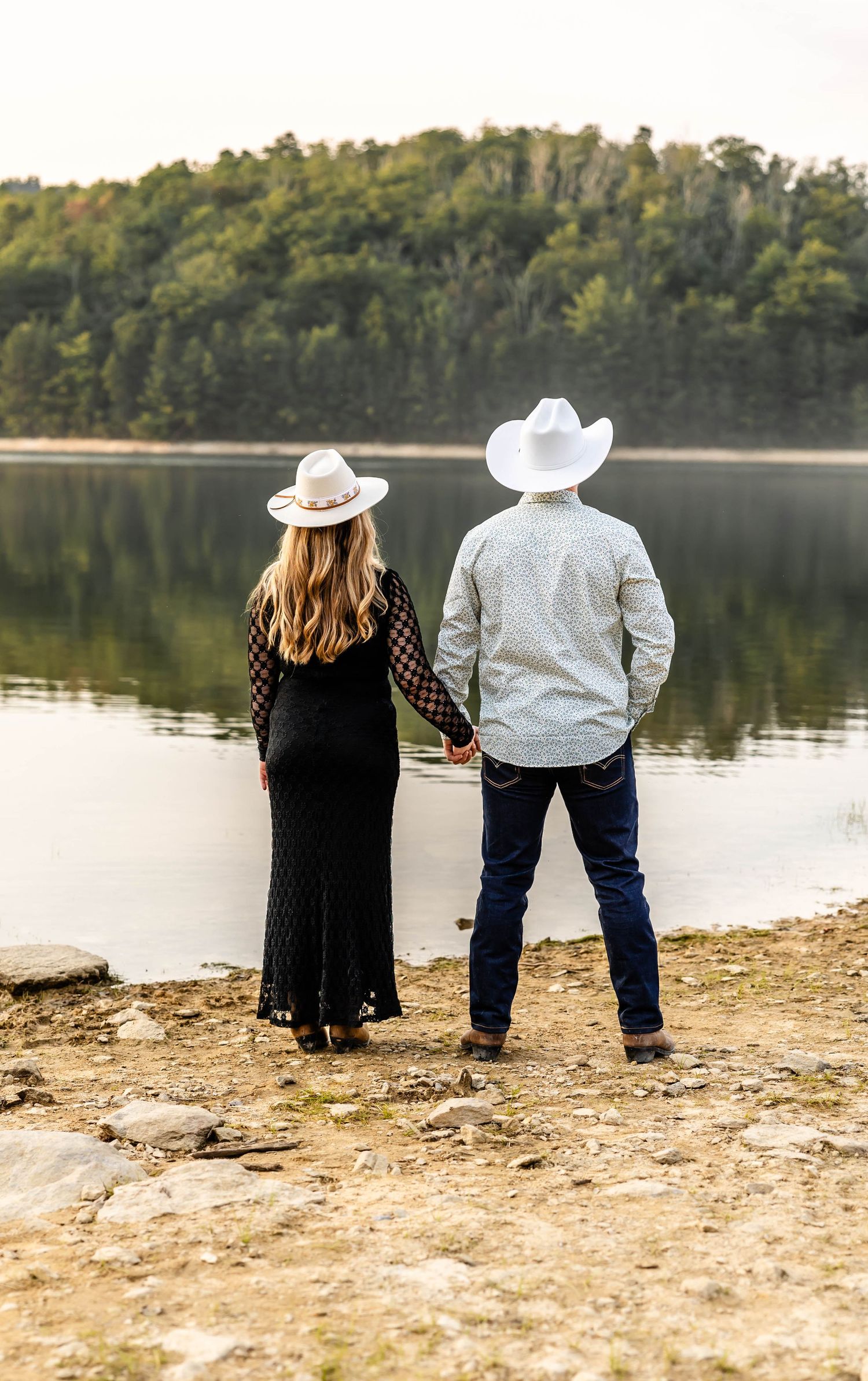 Couple in cowboy hats holds hands while looking out over a tranquil lake.