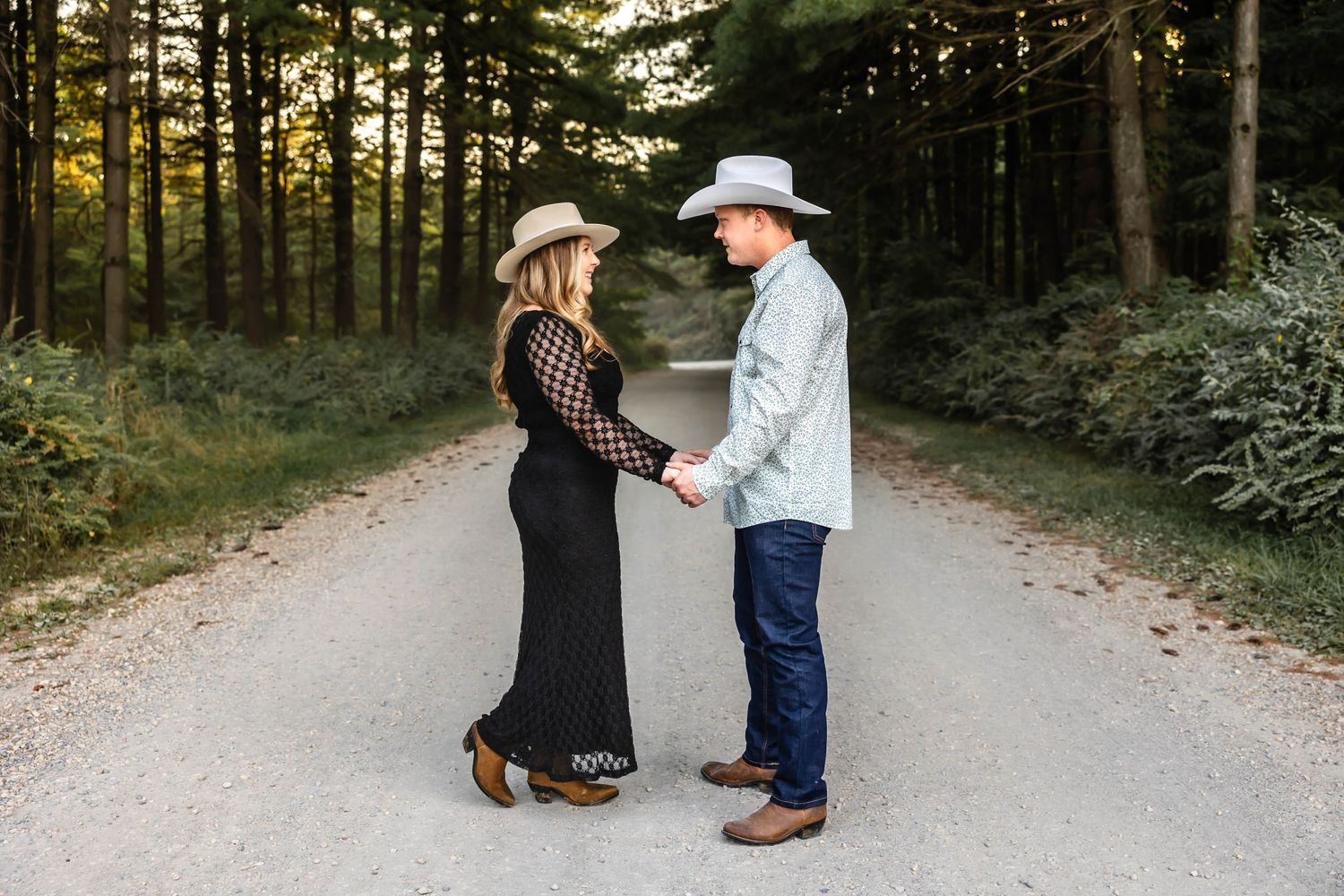 A couple in western attire holds hands while standing on a tree-lined dirt road.