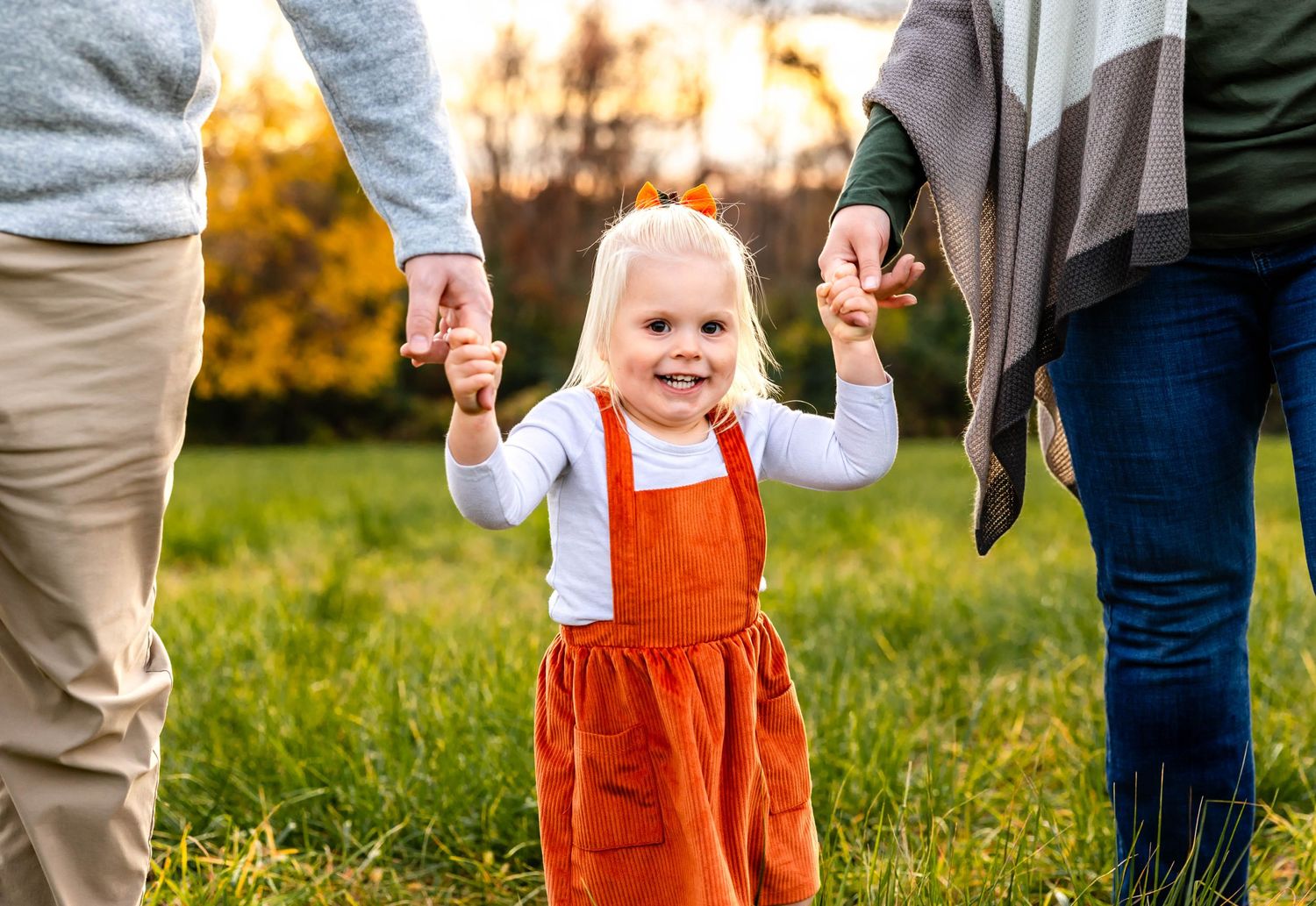 Joyful child in orange overalls holds hands with parents while walking outdoors at sunset.