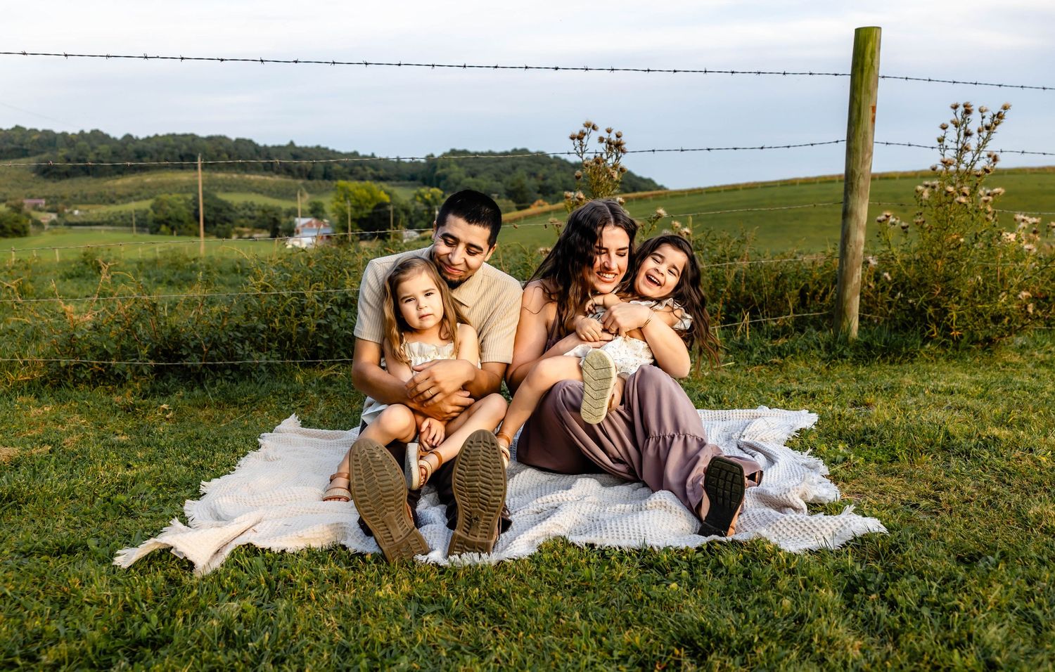 A family relaxes together on a white blanket in an open field with rolling hills in the background.